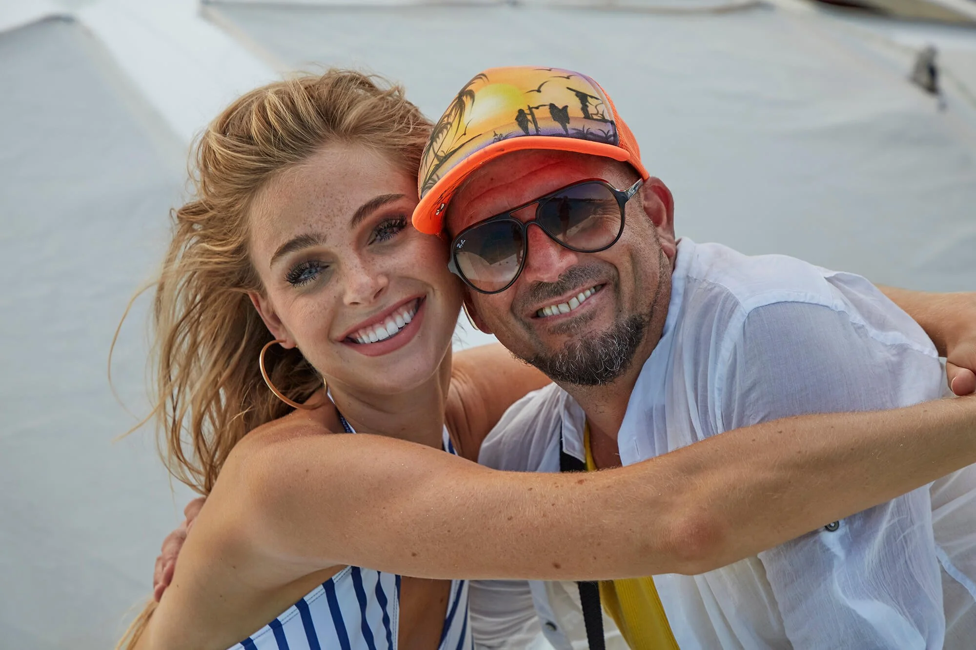 A smiling woman and man taking a selfie together on a boat with a body of water in the background. The woman has blonde hair and is wearing hoop earrings and a striped top. The man is wearing sunglasses, a colorful cap, and a white shirt.