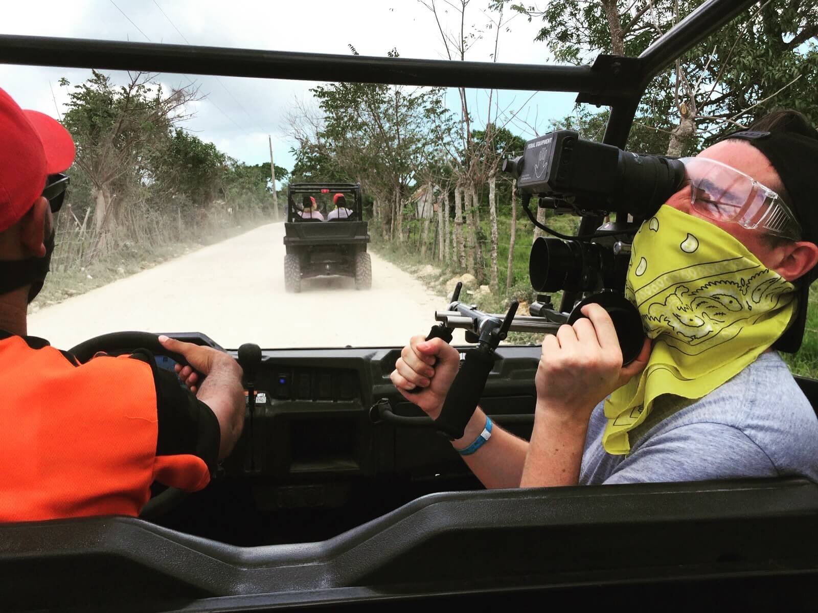 Person wearing glasses, a yellow bandana, and a gray shirt filming with a professional camera inside a utility vehicle, with another person steering at the front, on a dirt road lined with trees and a fence.