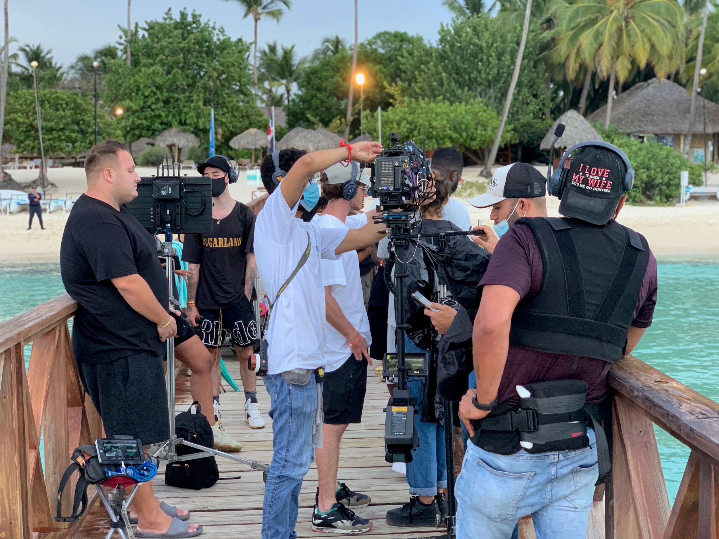 A film crew working on a shoot on a wooden dock by the beach, with palm trees and thatched-roof huts in the background, using cameras and audio equipment.