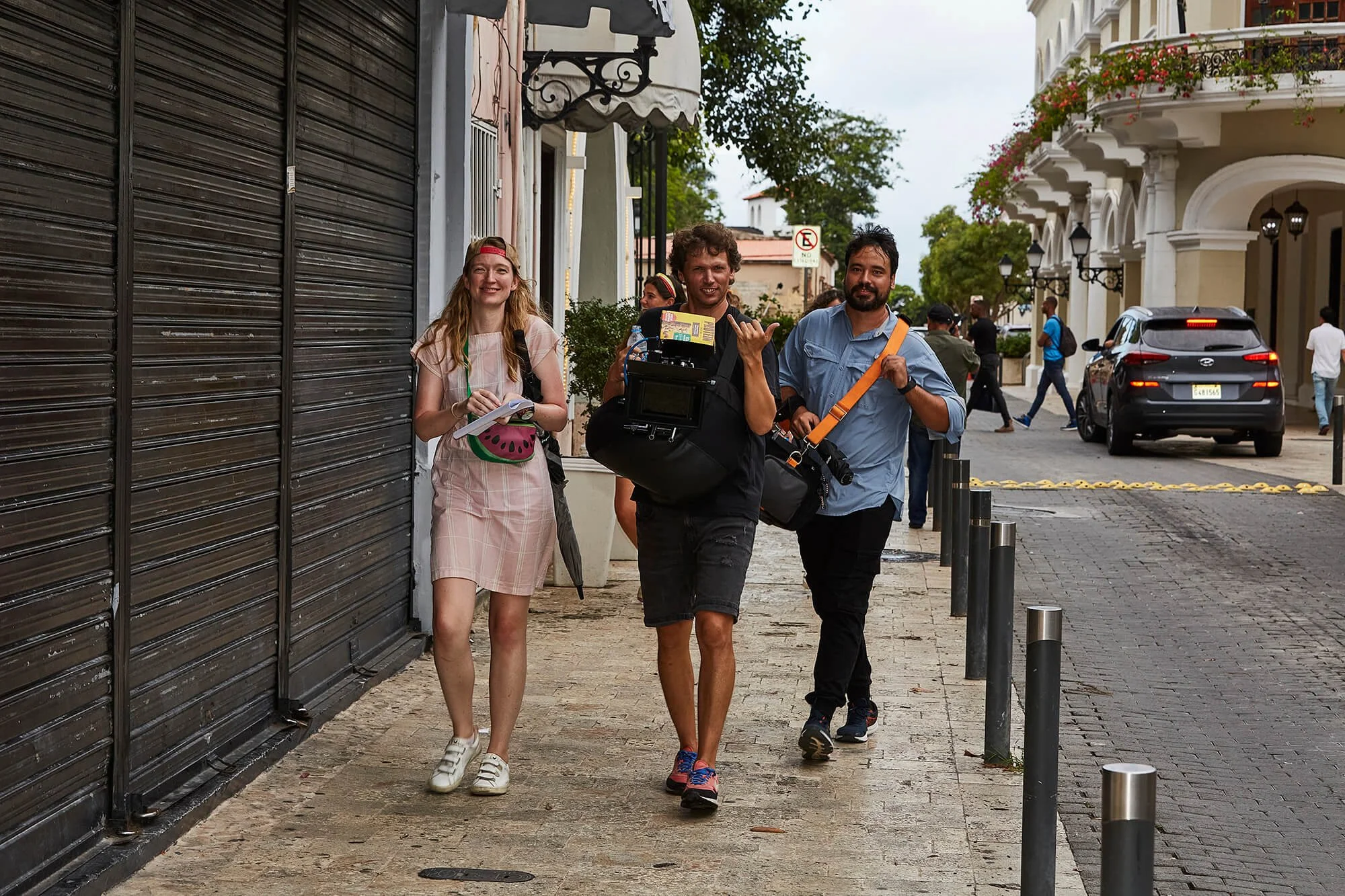 Group of three photographers walking on a city sidewalk, with a woman carrying a notebook and two men holding cameras and gear, on a cloudy day.