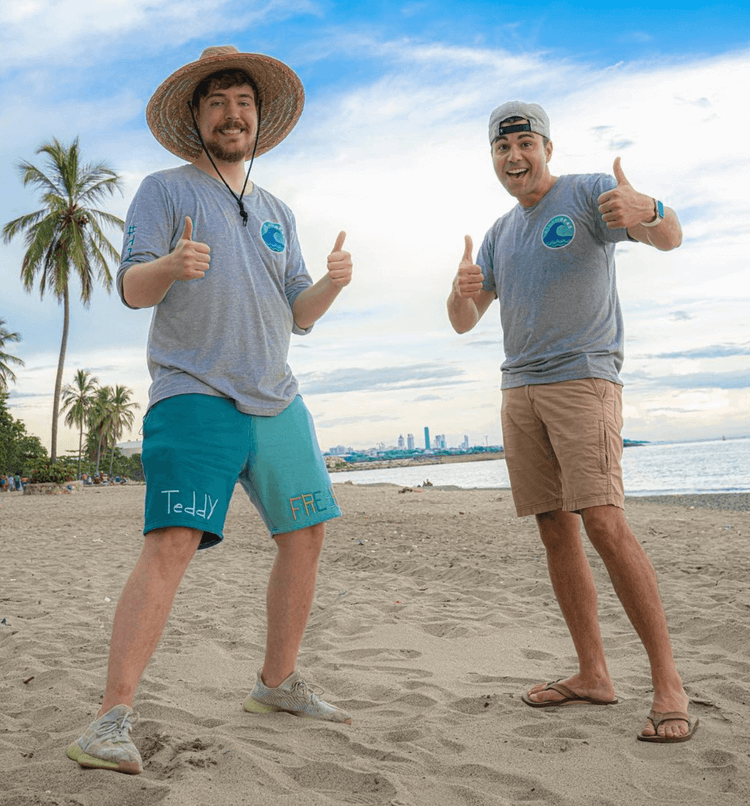 Two men standing on a sandy beach with palm trees, giving thumbs up and smiling, with city skyline in the background on a partly cloudy day. They are wearing matching gray t-shirts with a wave logo.