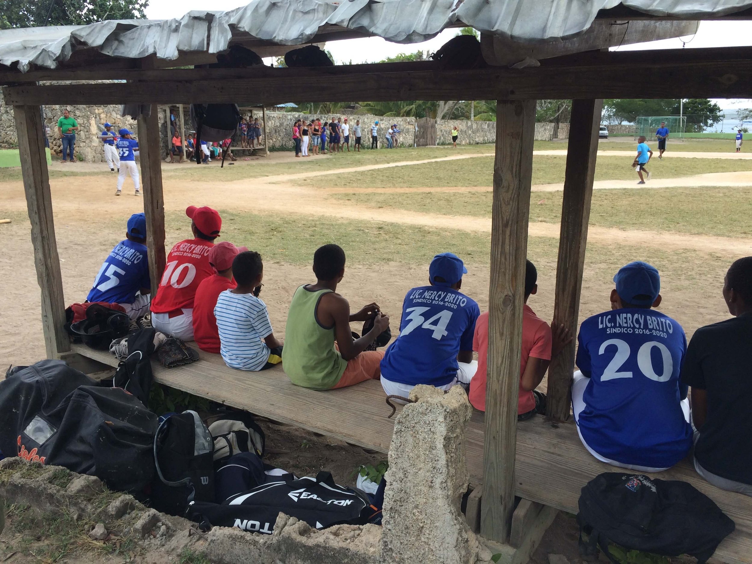 Children and people watching a baseball game under a wooden shelter on a grassy field.