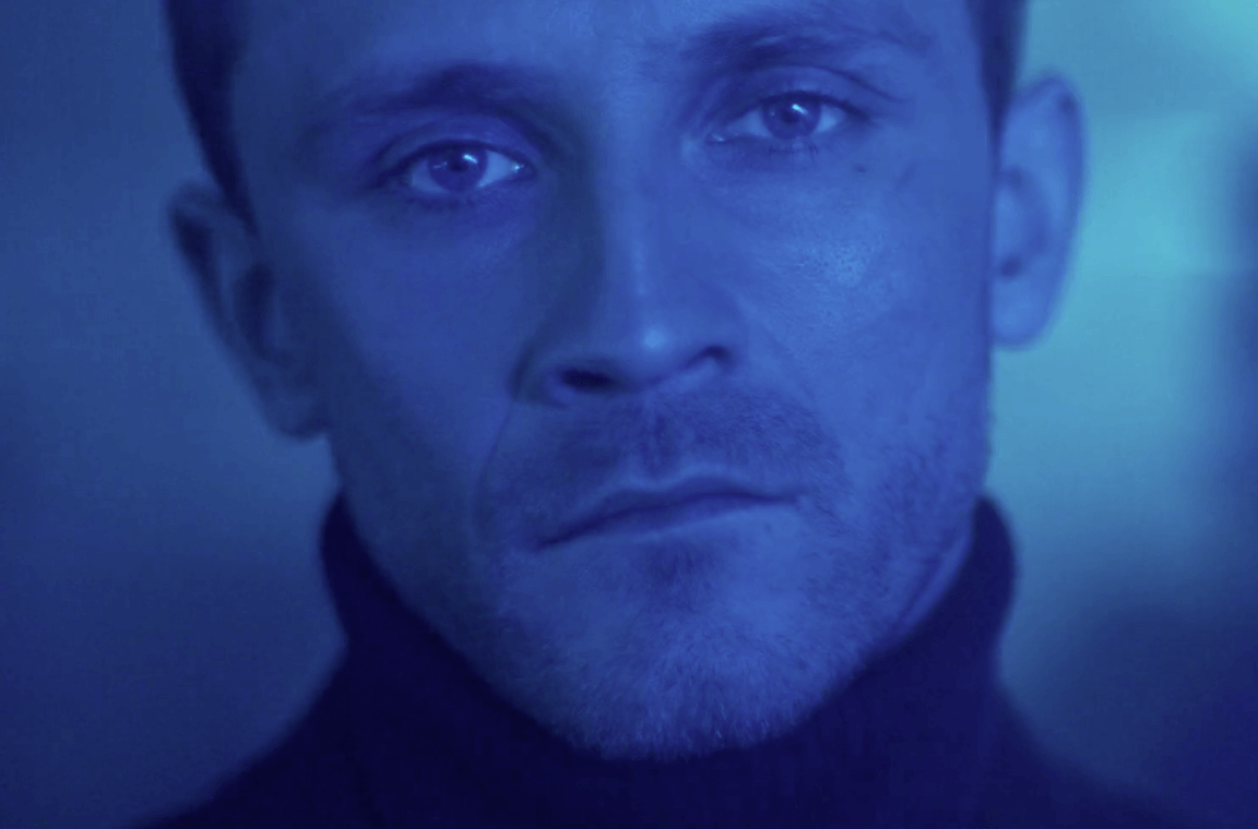 Close-up of a young man's face with blue lighting, wearing a black shirt or jacket.