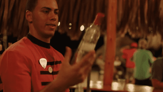 A young man wearing a red and black striped shirt with a name tag on it, looks at a clear bottle, inside a busy outdoor dining area with strings of lights and other people in the background.