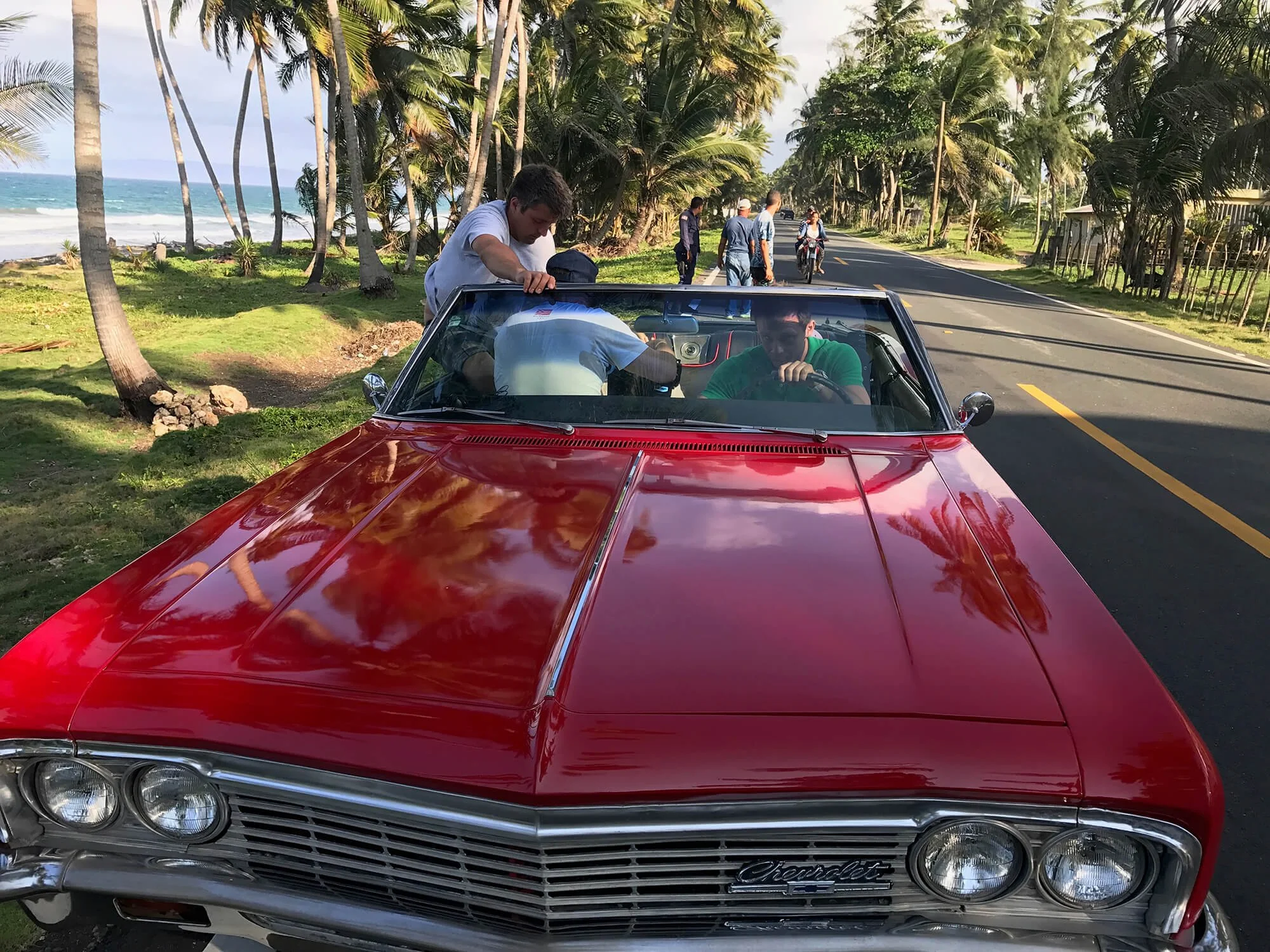 A red vintage Chevrolet convertible car parked on the side of a road by a beach with palm trees. Several people are around the car, with two men sitting inside, one in the driver's seat and the other in the passenger seat, and others standing near th