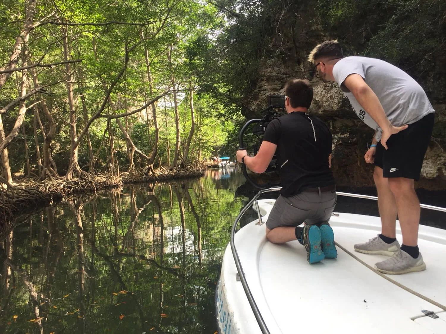 Two men on a boat in a lush, green mangrove forest, with one kneeling and operating a camera while the other stands nearby.