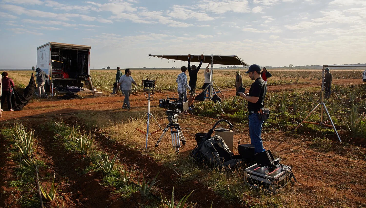 A film crew setting up equipment in a field for an outdoor shoot with cameras, lighting, and a crew member holding a script or notes.