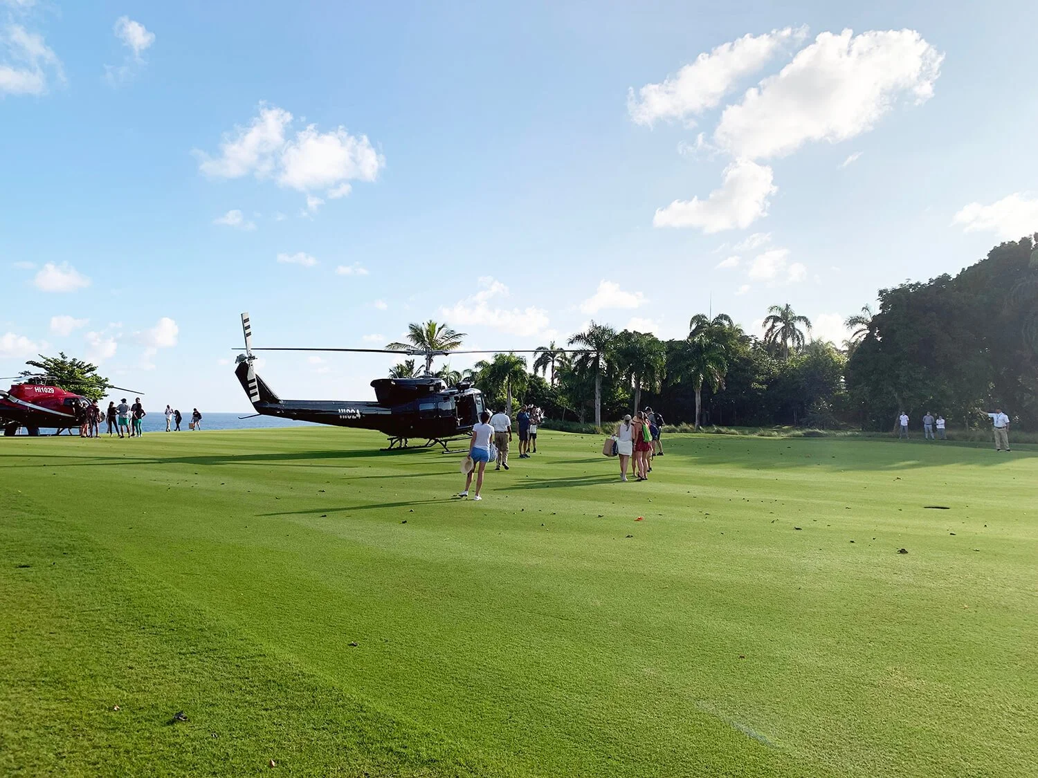 A helicopter on a lush green golf course with people walking and standing, some near the helicopter and others at a distance, under a blue sky with scattered clouds and palm trees in the background.
