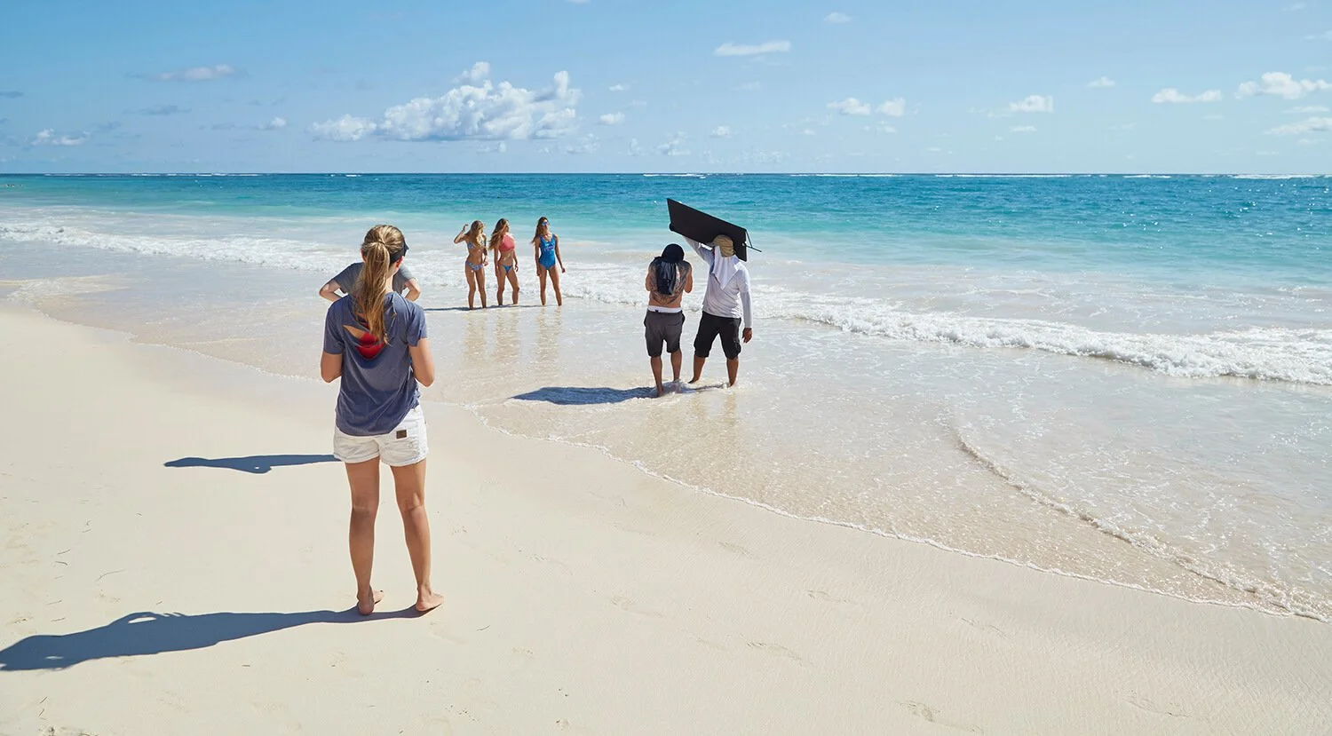People on a beach taking photos of four women in swimsuits near the shoreline, with a woman in the foreground observing.
