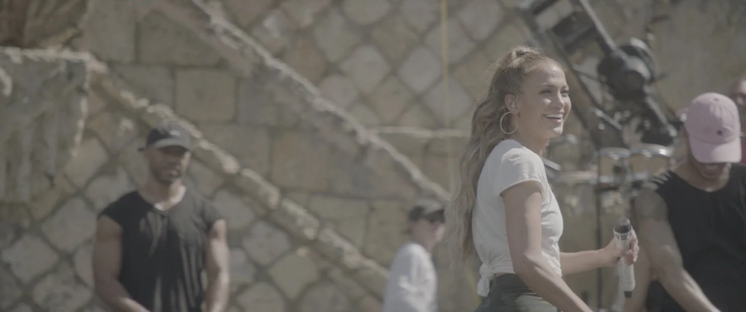 Smiling woman with long wavy hair holding a microphone at outdoor event with stone wall and people in background.
