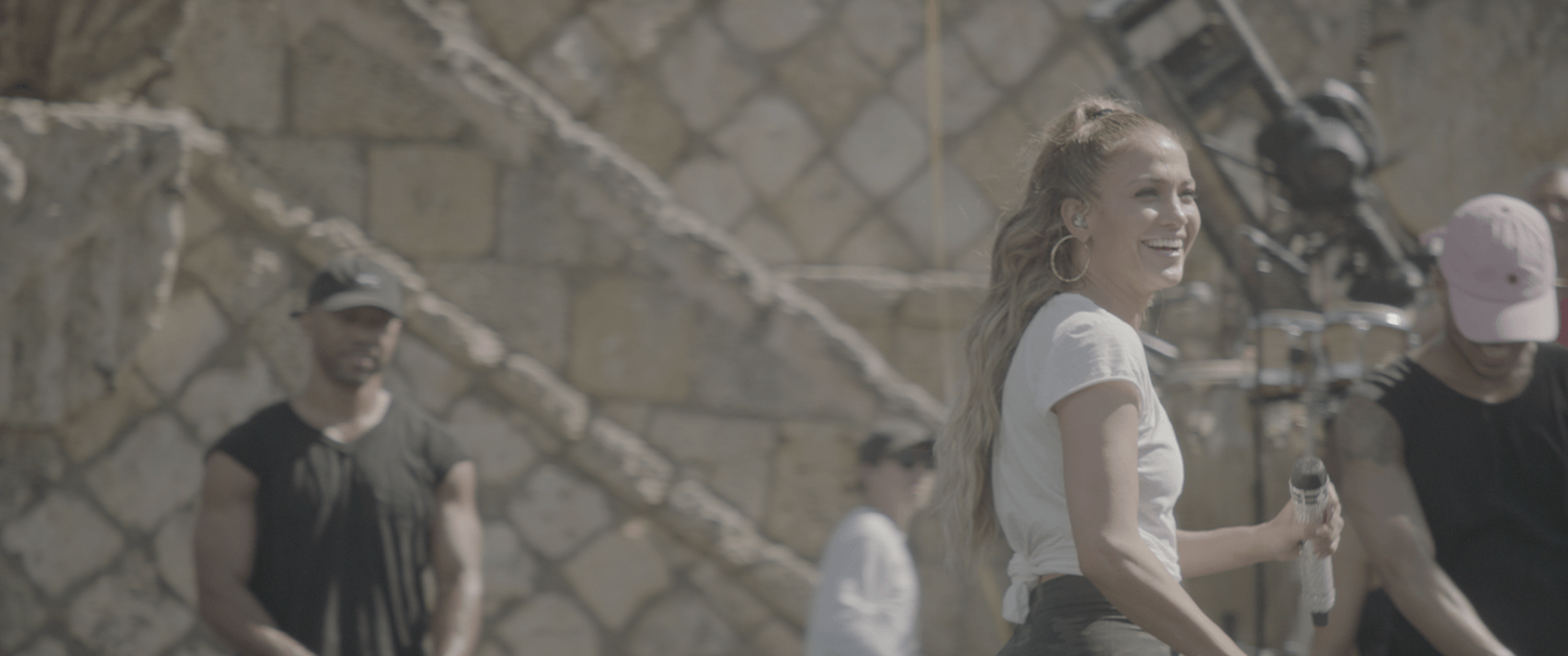 A woman smiling, holding a microphone, with other people and a stone wall in the background.