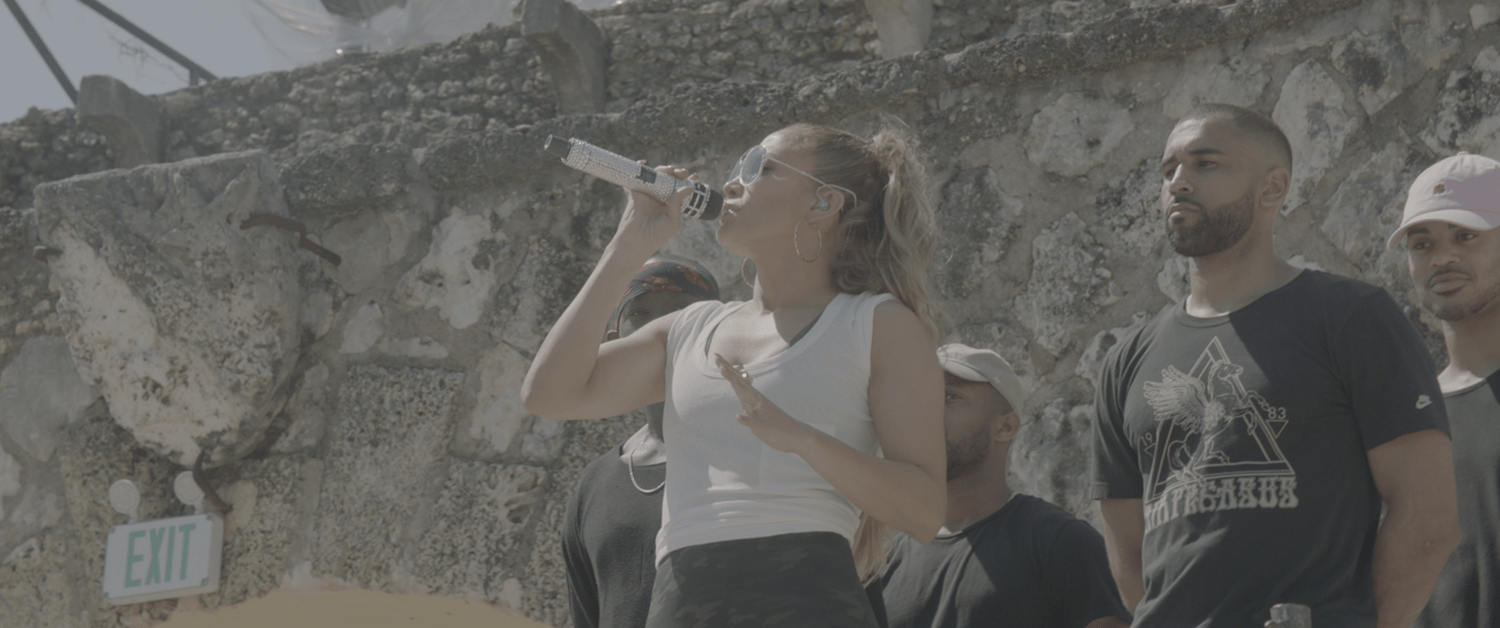 A woman with sunglasses and hoop earrings singing into a microphone, standing outdoors against a rocky wall, with three men standing nearby, some with serious expressions.