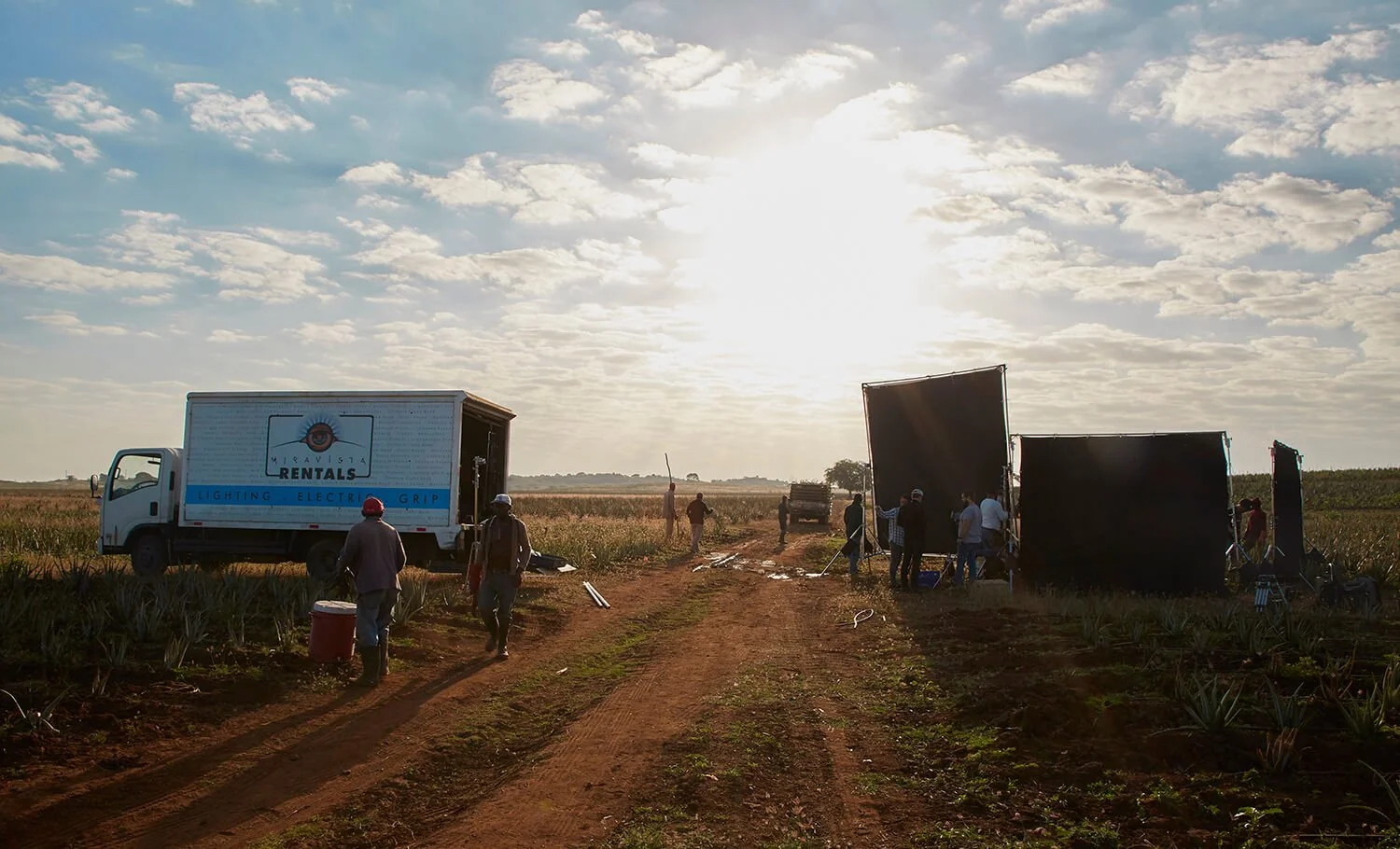 Film crew setting up equipment in an agricultural field during sunset, with a truck labeled 'Miqvista Rentals' and large black panels for filming.