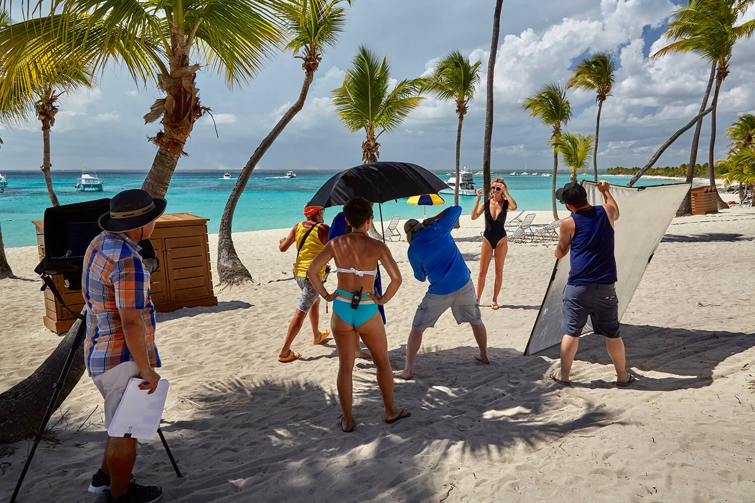 A beach photoshoot in progress with a woman in a black swimsuit posing, surrounded by a crew including a photographer, an assistant holding a reflector, and others adjusting equipment, under palm trees by the ocean.