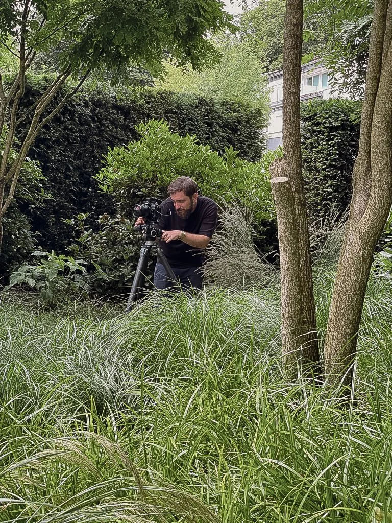 Ein Mann mit Kamera in einem dichten, grünen Garten mit Büschen, Bäumen und hohem Gras.