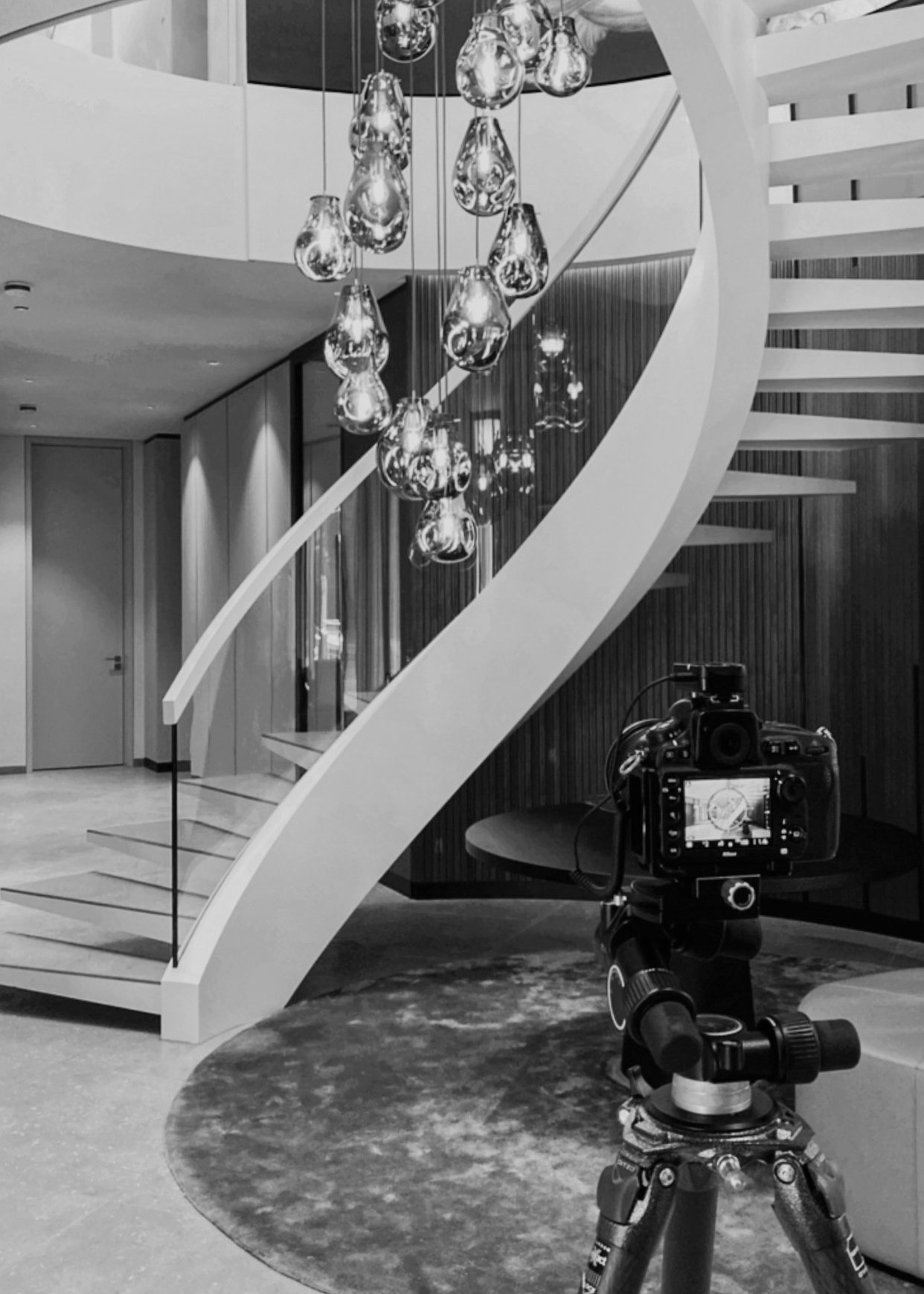 Innenaufnahme eines modernen Hauses mit eleganter Treppe, einer speziellen Hängelampe, einer Kamera auf einem Stativ im Vordergrund und einer Sitzgelegenheit im Hintergrund.