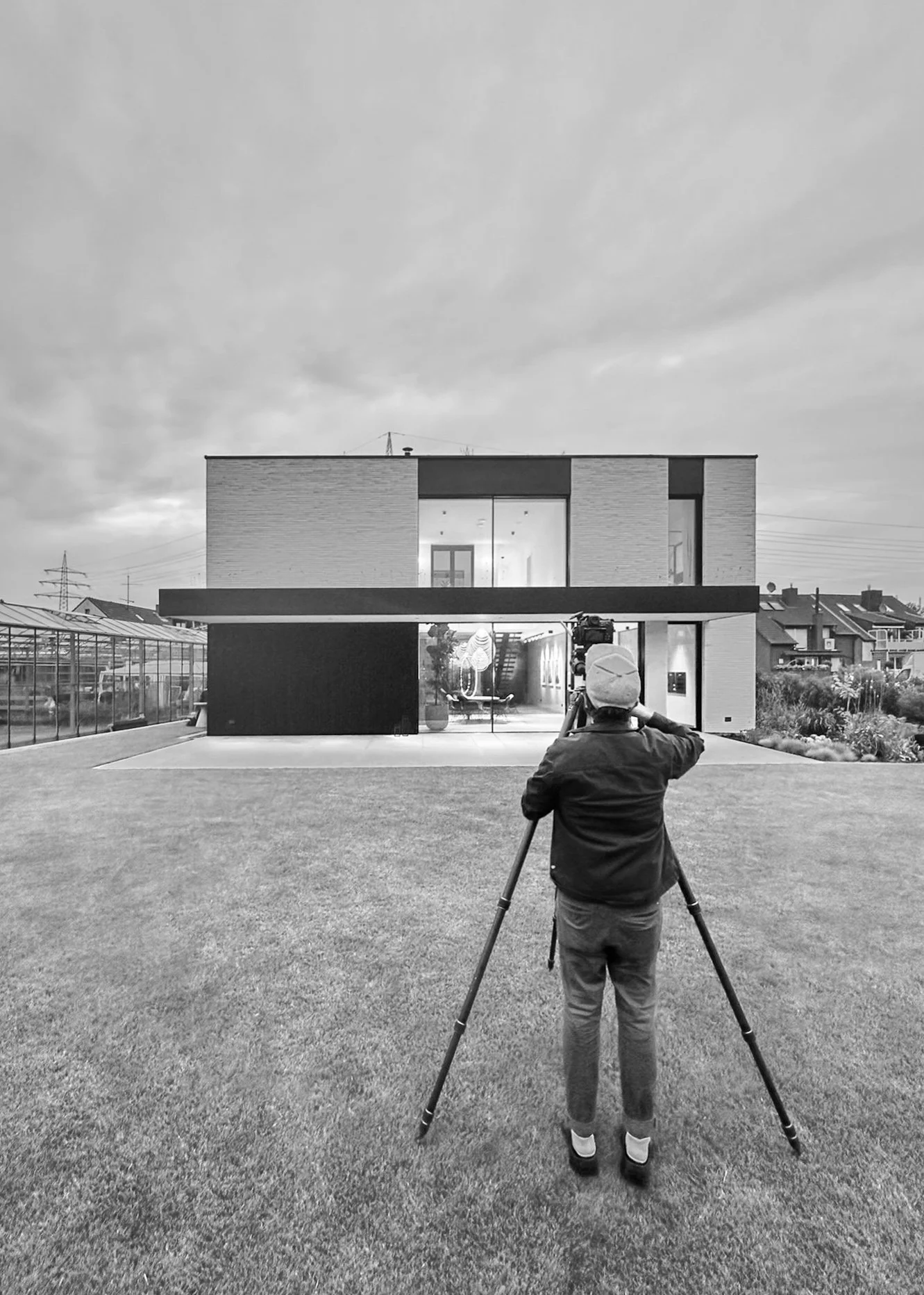 Ein Fotograf fotografiert ein modernes Haus mit einem Fernglas auf einem Stativ auf einer grünen Wiese, im Hintergrund ein bewölkter Himmel.
