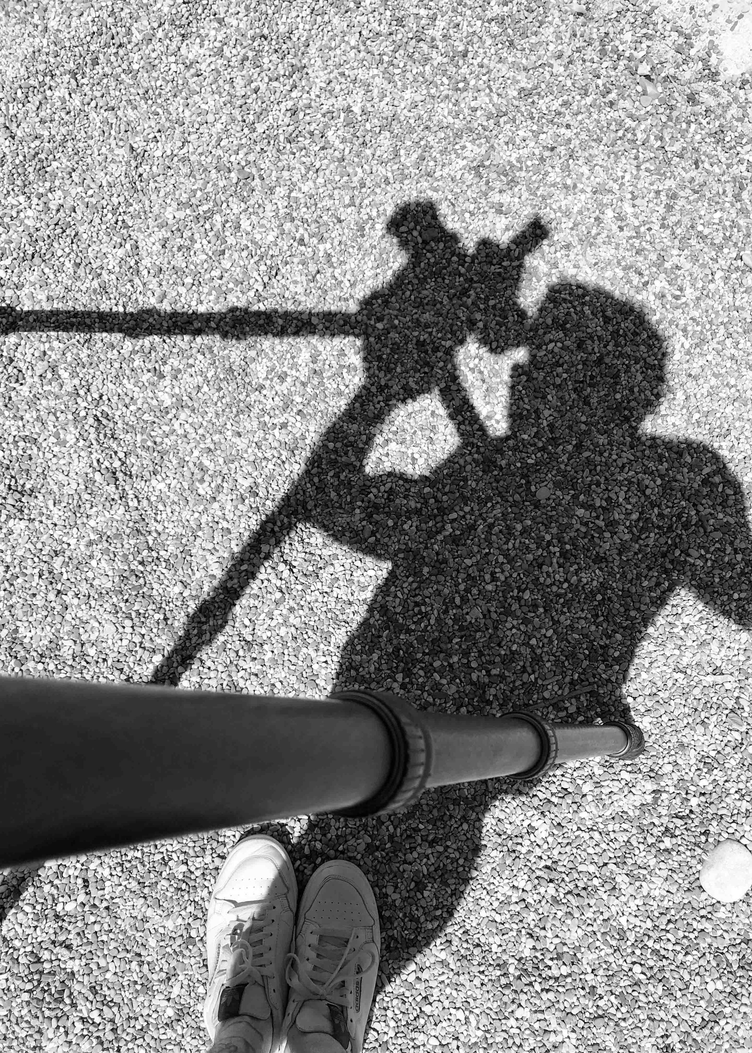 Schattenspiel eines Menschen, der eine Blume hält, auf einem sandigen Boden, aufgenommen aus der Perspektive der Person, die die Foto macht.