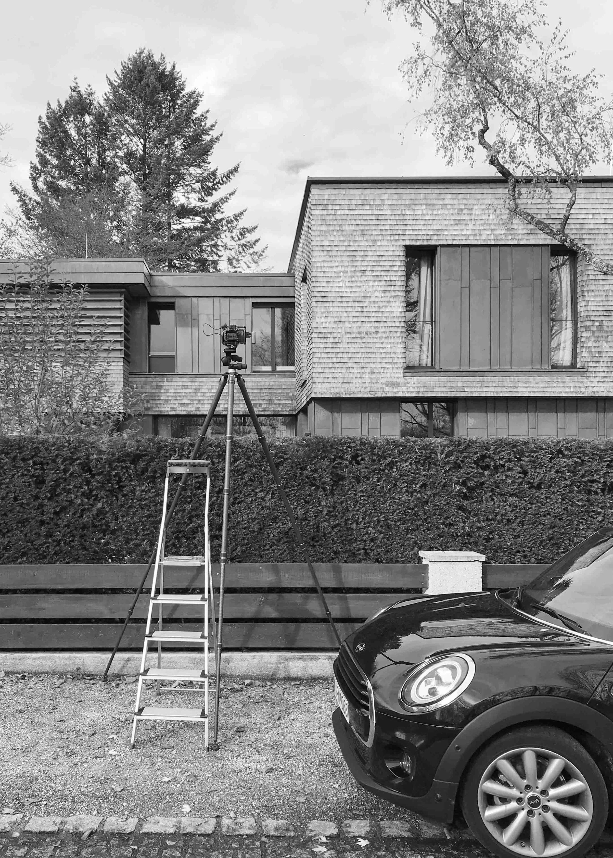 Ein Haus mit moderner Architektur, ein Fernauslöser auf einem Stativ, ein Auto und ein Gehweg davor. Der Hintergrund zeigt Bäume und Wolken.