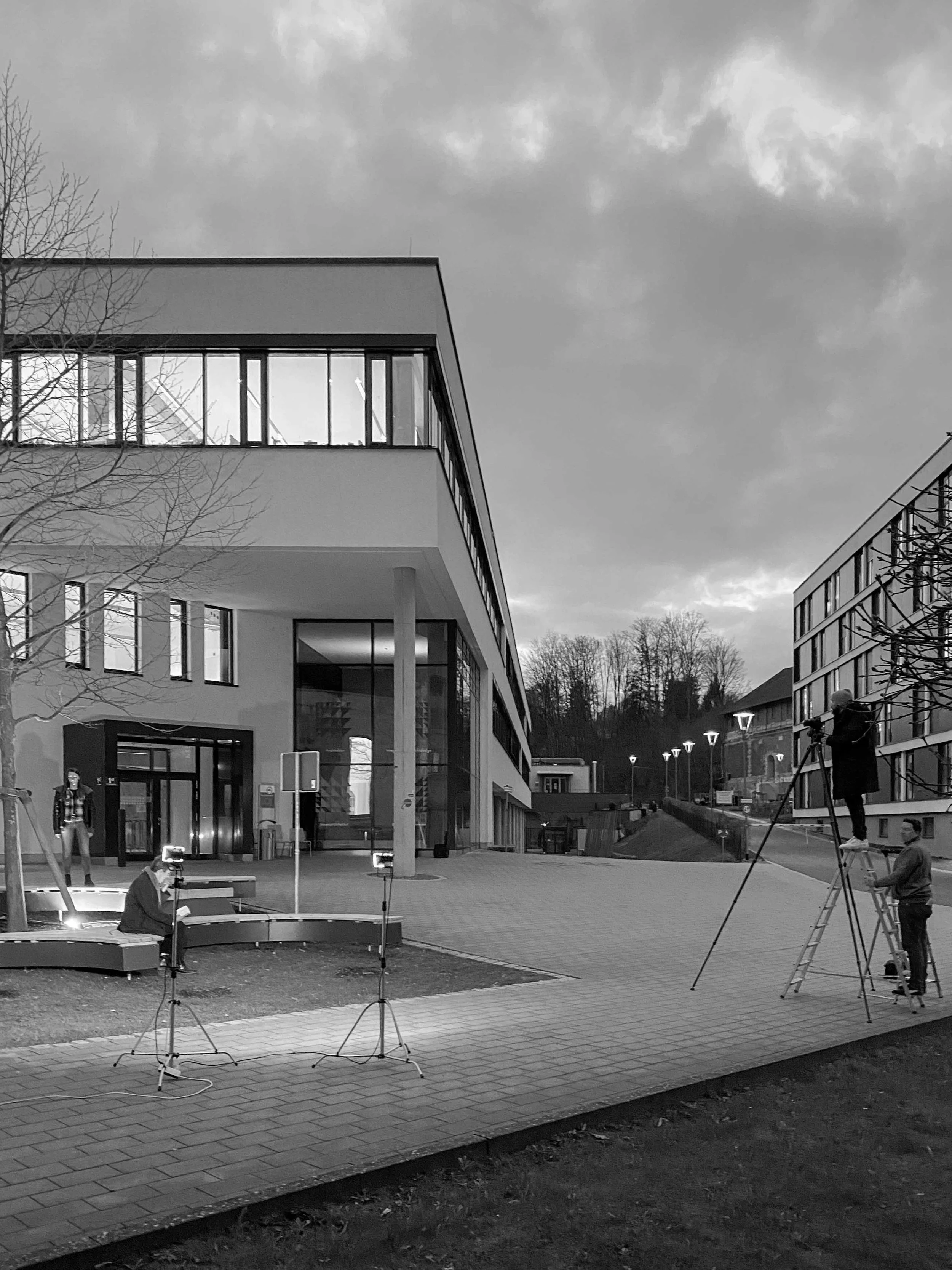 Drei Film- oder Fernsehkameras werden auf ein modernes Gebäude gerichtet, während eine Person auf einem Leitern steht, um die Aufnahme zu steuern. Im Vordergrund sitzt eine Person, die möglicherweise den Ton überprüft. Es ist Abend oder bewölktes Wetter.