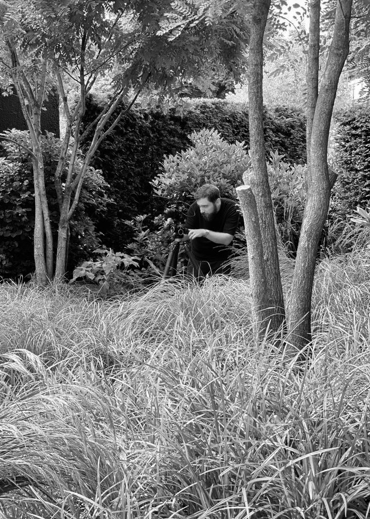 Ein Mann mit dunklem Haar und Bart, der mit einer Kamera auf einem Stativ im Garten fotografiert, umgeben von Büschen und Bäumen.