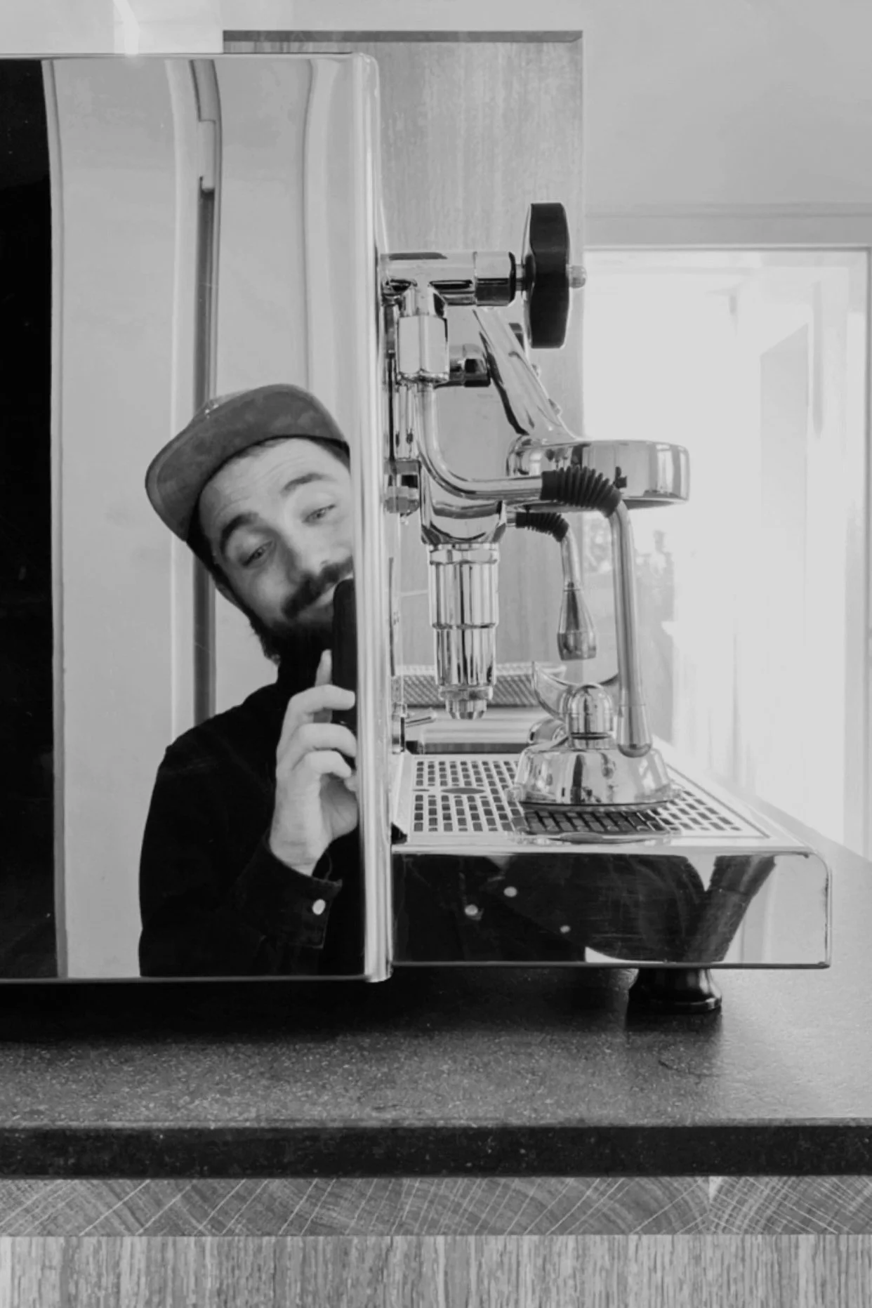 Schwarz-weiß-Foto eines Mannes, der sich hinter einer Espressomaschine versteckt und in den Spiegel schaut, während er eine Tasse haltend lächelt.