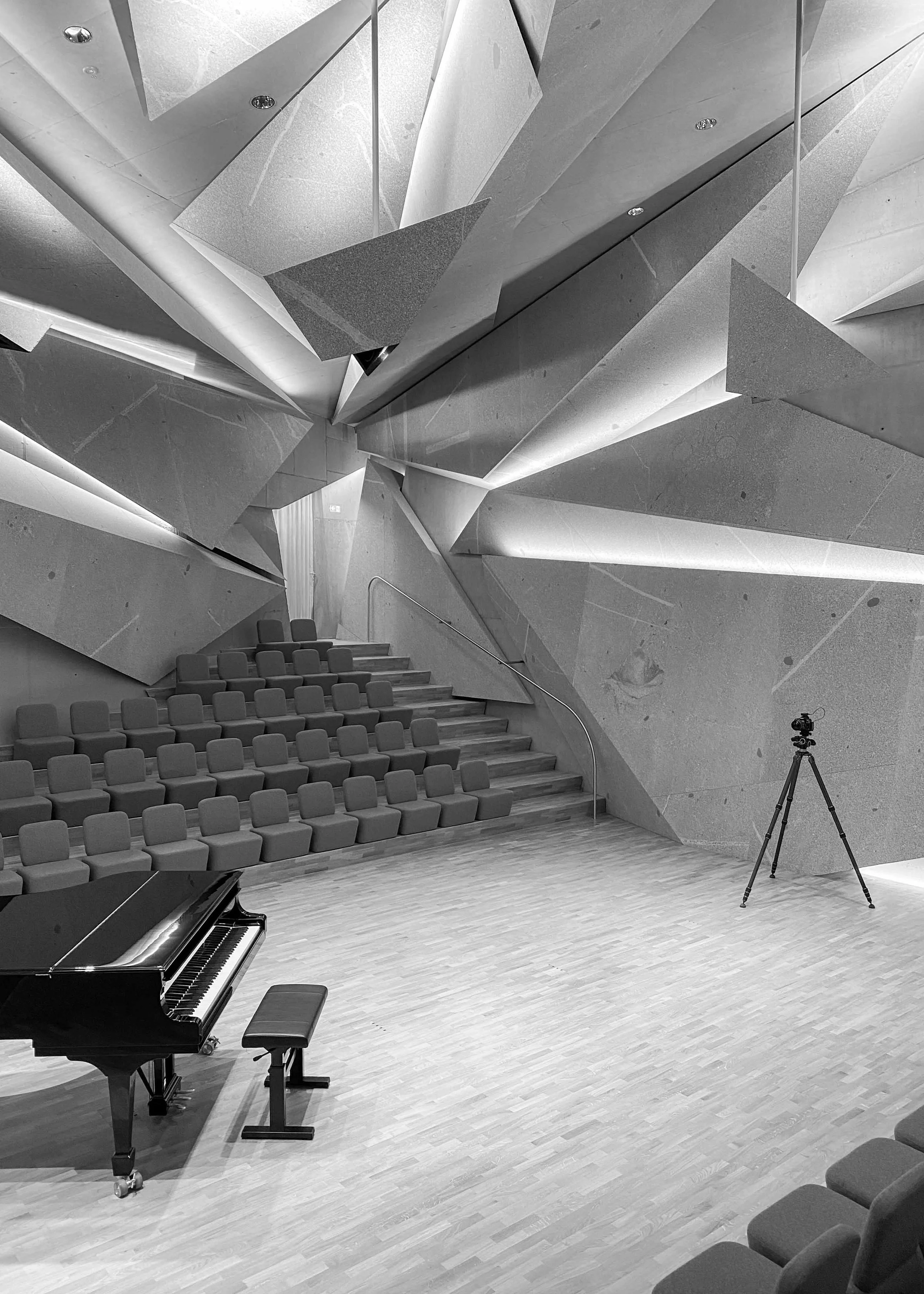 Ein moderner Konzertsaal mit leuchtender, geometrischer Wandgestaltung, Sitzreihen, einem Flügel, einer Kamera auf einem Stativ und einer kleinen Treppe.