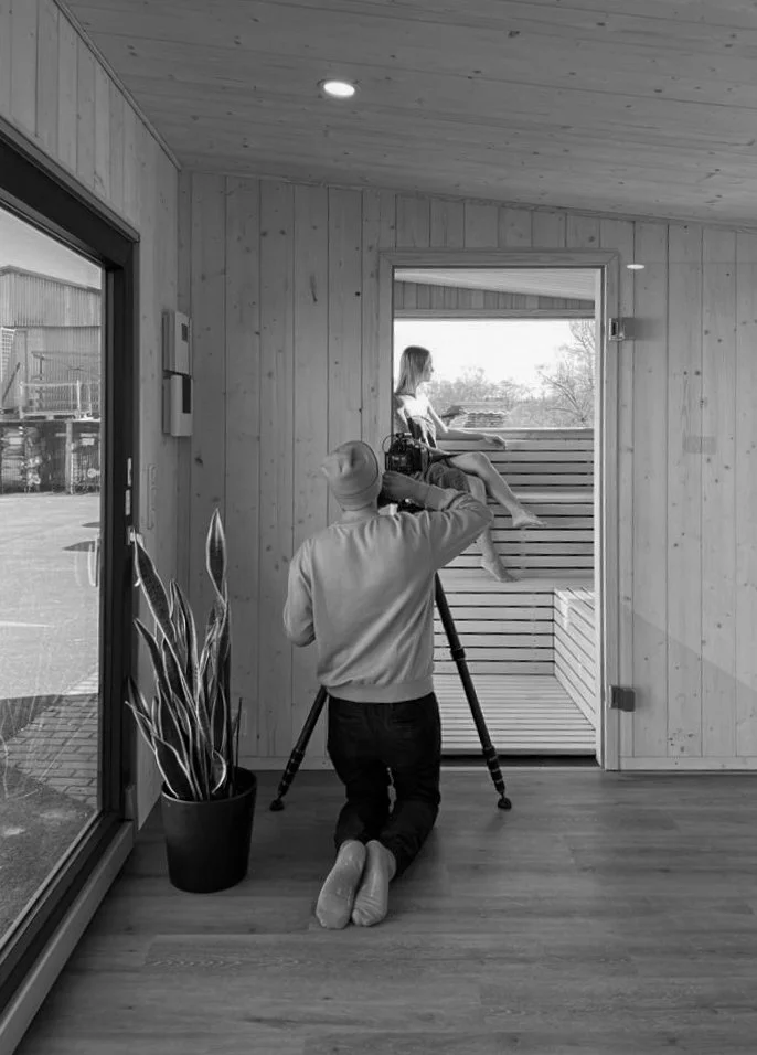Ein Fotograf filmt eine Frau, die auf einer Bank draußen auf einer Veranda sitzt, durch ein Fenster. Das Bild ist in Schwarz-Weiß.