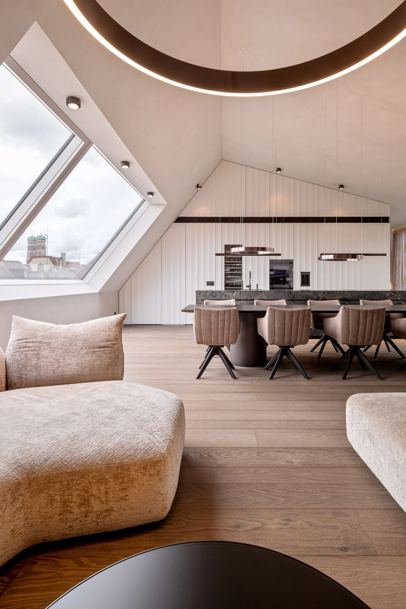 Moderner, heller Essbereich mit runder Tisch, gesteppten Stühlen, offener Küche im Hintergrund, große Dachfenster mit Stadtblick, minimalistische Decke mit runden Beleuchtungskörpern, Holzfußboden.