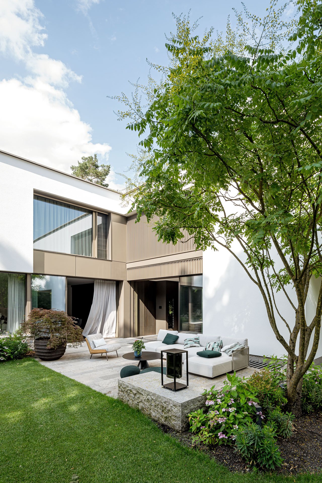 Moderne Terrasse mit Gartenmöbeln, umgeben von grünen Pflanzen und einem großen Baum, vor einem weißen Haus mit großen Fenstern.