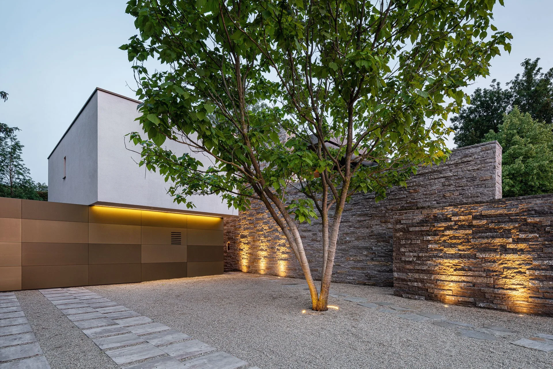 Moderne Gartenanlage mit Sandboden, Kiesweg, Baum und einer weißen Hausfassade mit goldener Beleuchtung bei Dämmerung.