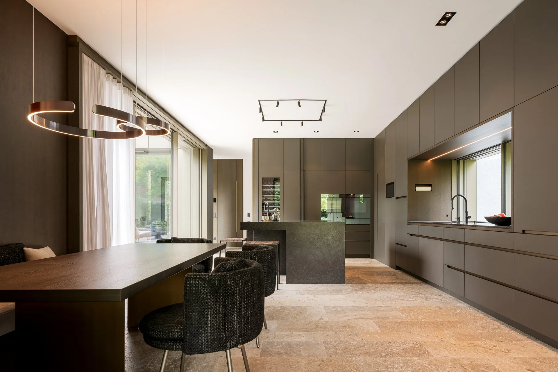 Moderne, offene Küche und Essbereich mit dunklen und neutralen Farbtönen, einem langen Esstisch mit schwarzen Stühlen, einer dunklen Kücheninsel, großen Fenstern mit Vorhängen, und minimalistischer Beleuchtung.