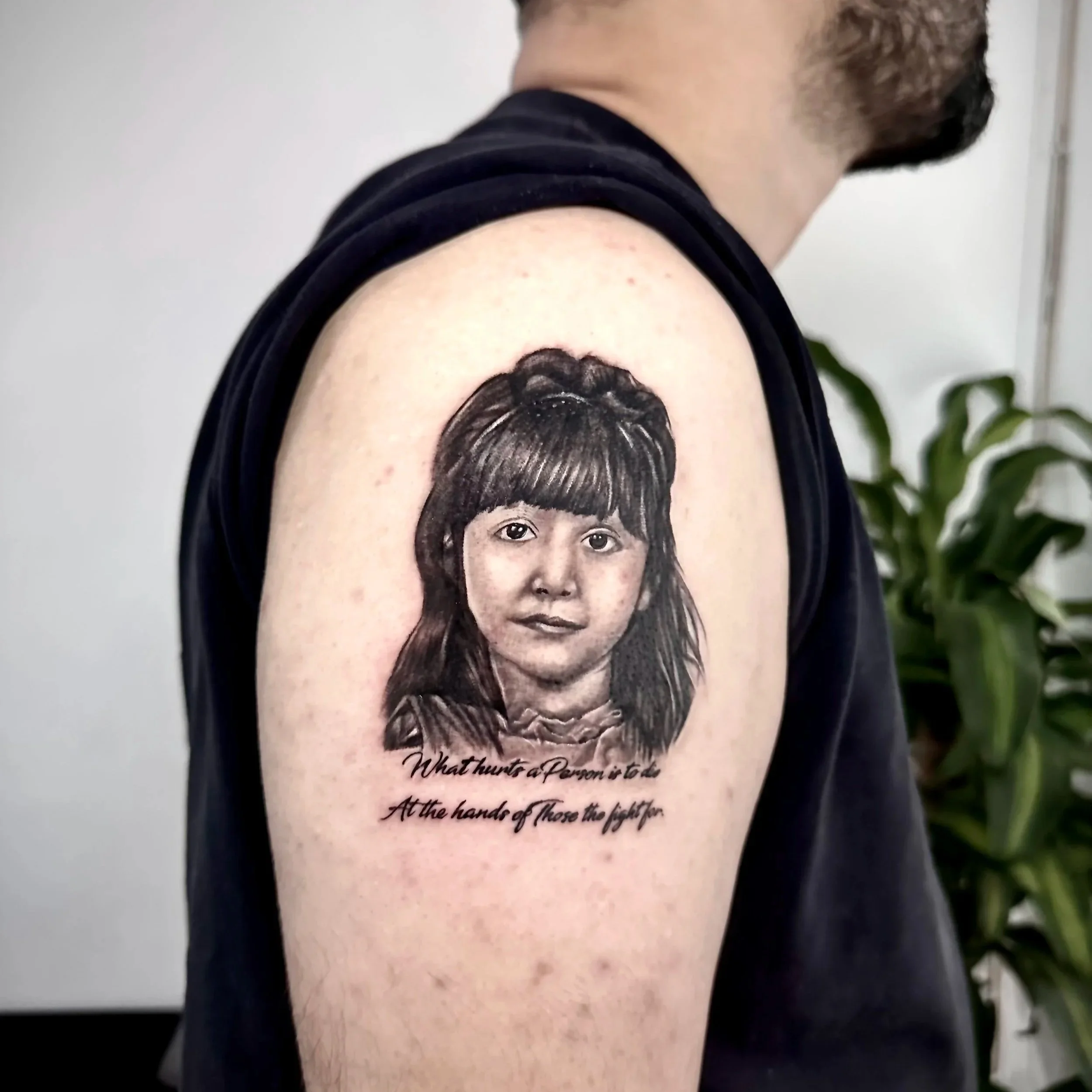 A tattoo on a person's upper arm depicting a young girl with dark hair and bangs, accompanied by a quote that reads, 'What hinders a person is not the attack but the fight.'