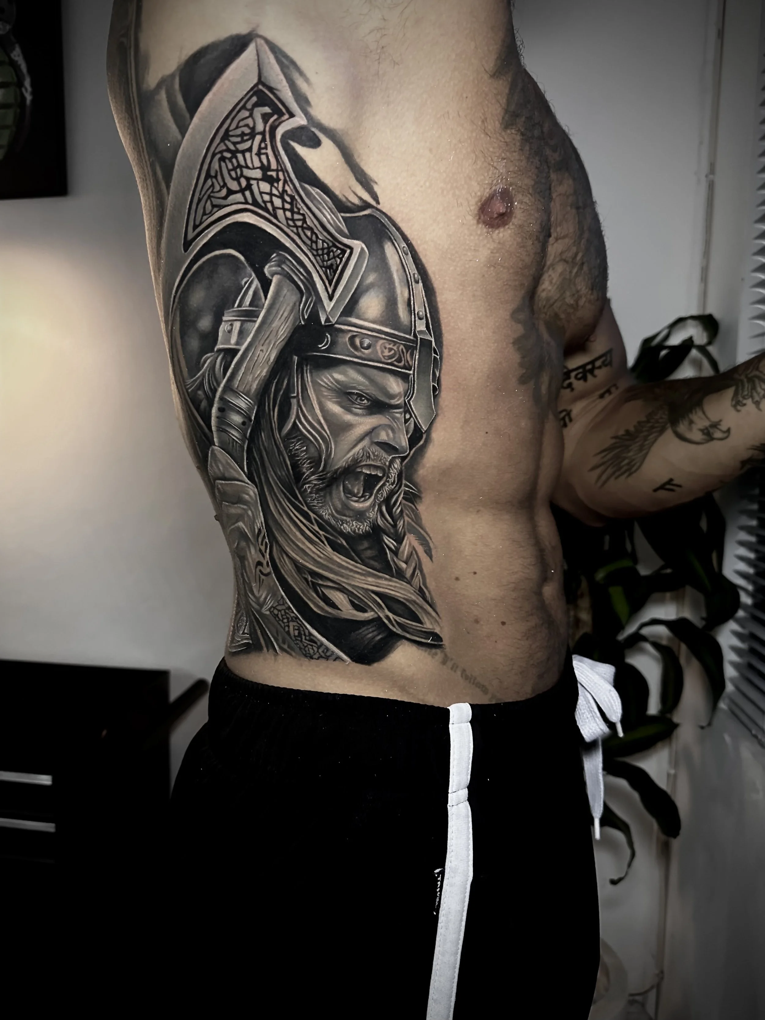 A detailed black and gray tattoo of a fierce warrior wearing a helmet and armor, depicting a bearded man with a yelling expression, on a person's left upper arm and shoulder.
