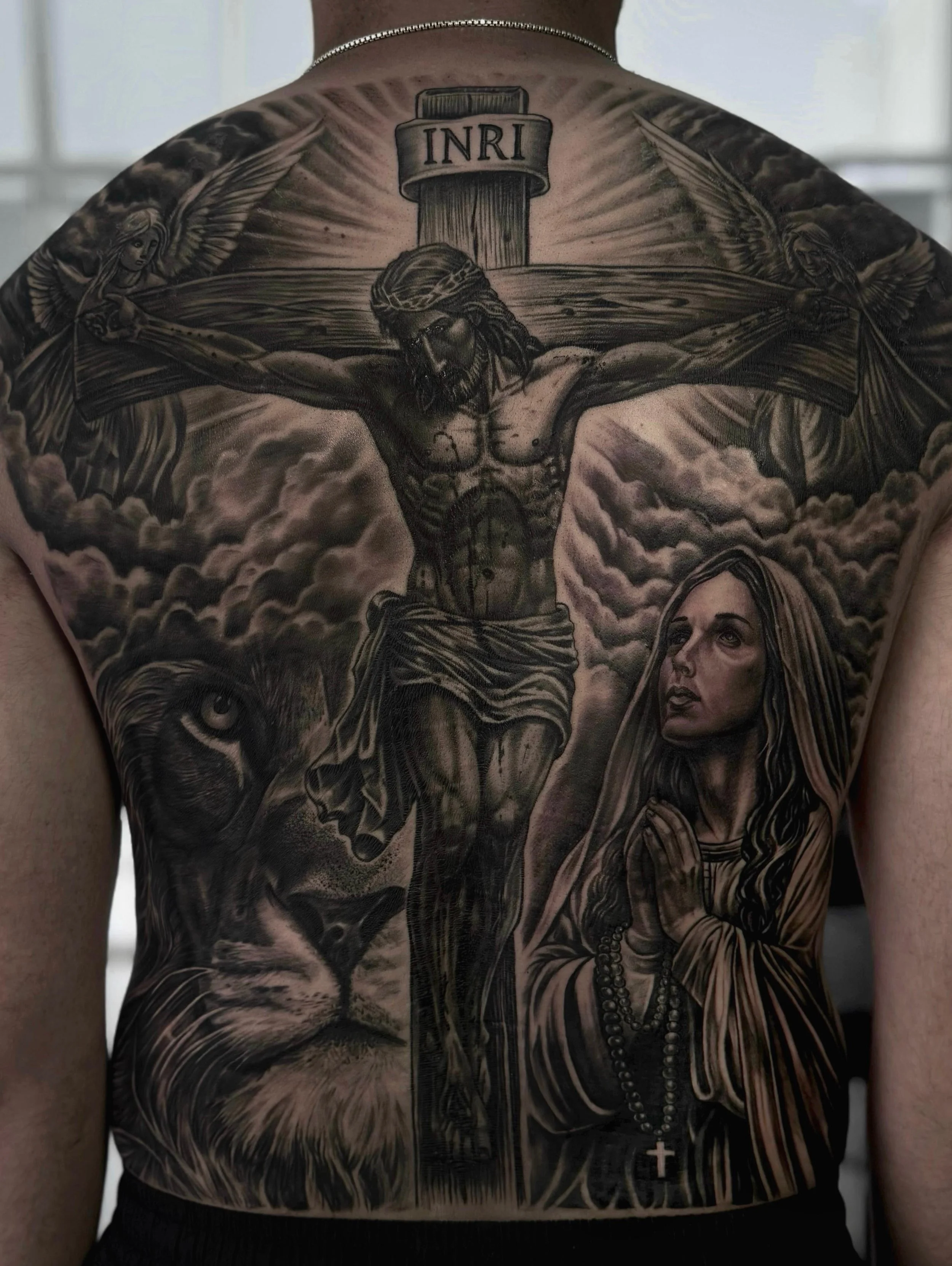 A black and gray tattoo on a person's back depicts a crucifixion scene with Jesus Christ on the cross, flanked by angels, with clouds below and a Bible with 'INRI' above the cross. A woman with a rosary and a cross pendant is at the bottom right of t