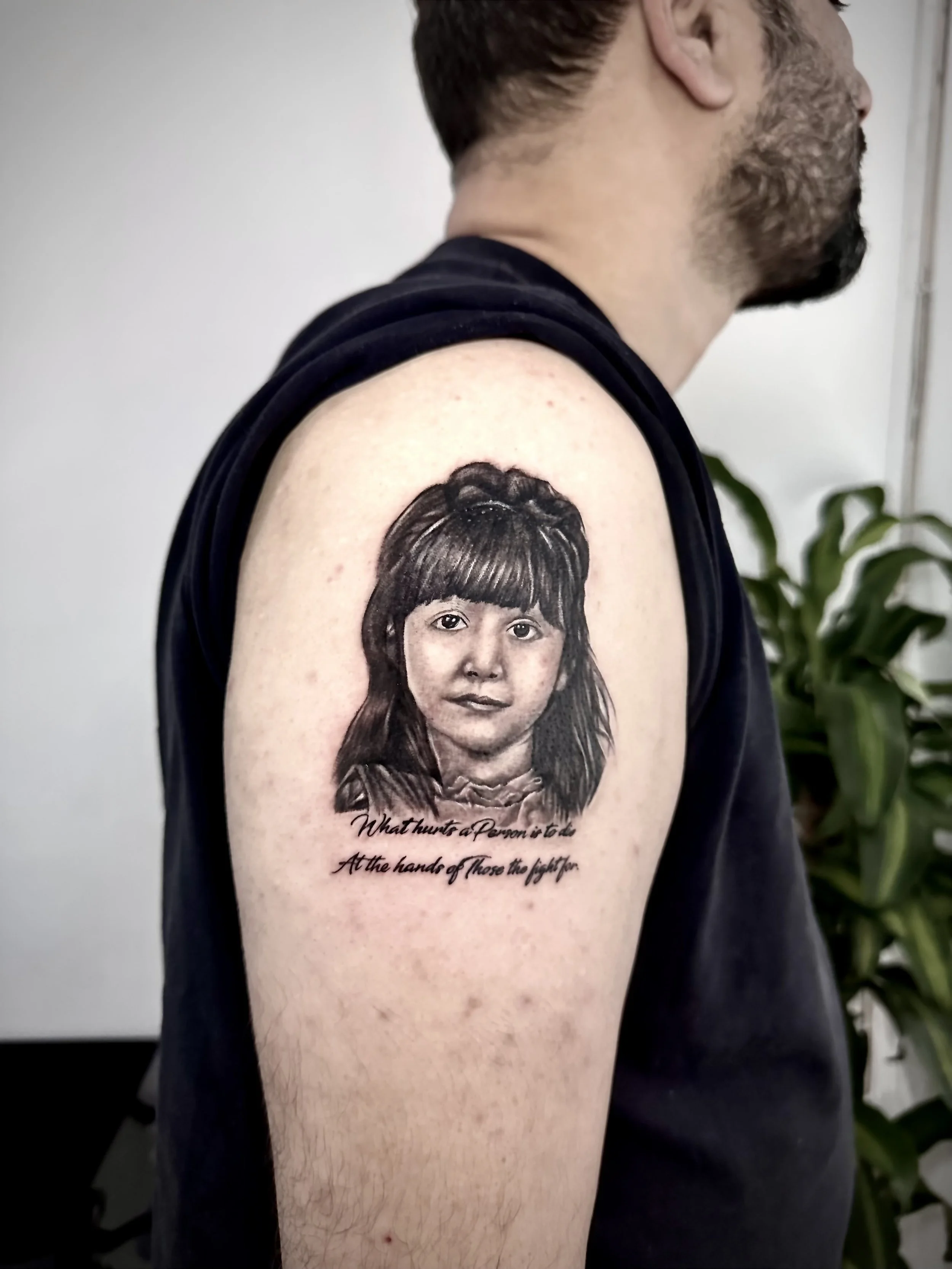 A tattoo on a person's upper arm depicting a young girl with dark hair and bangs, accompanied by a quote that reads, 'What hinders a person is not the attack but the fight.'