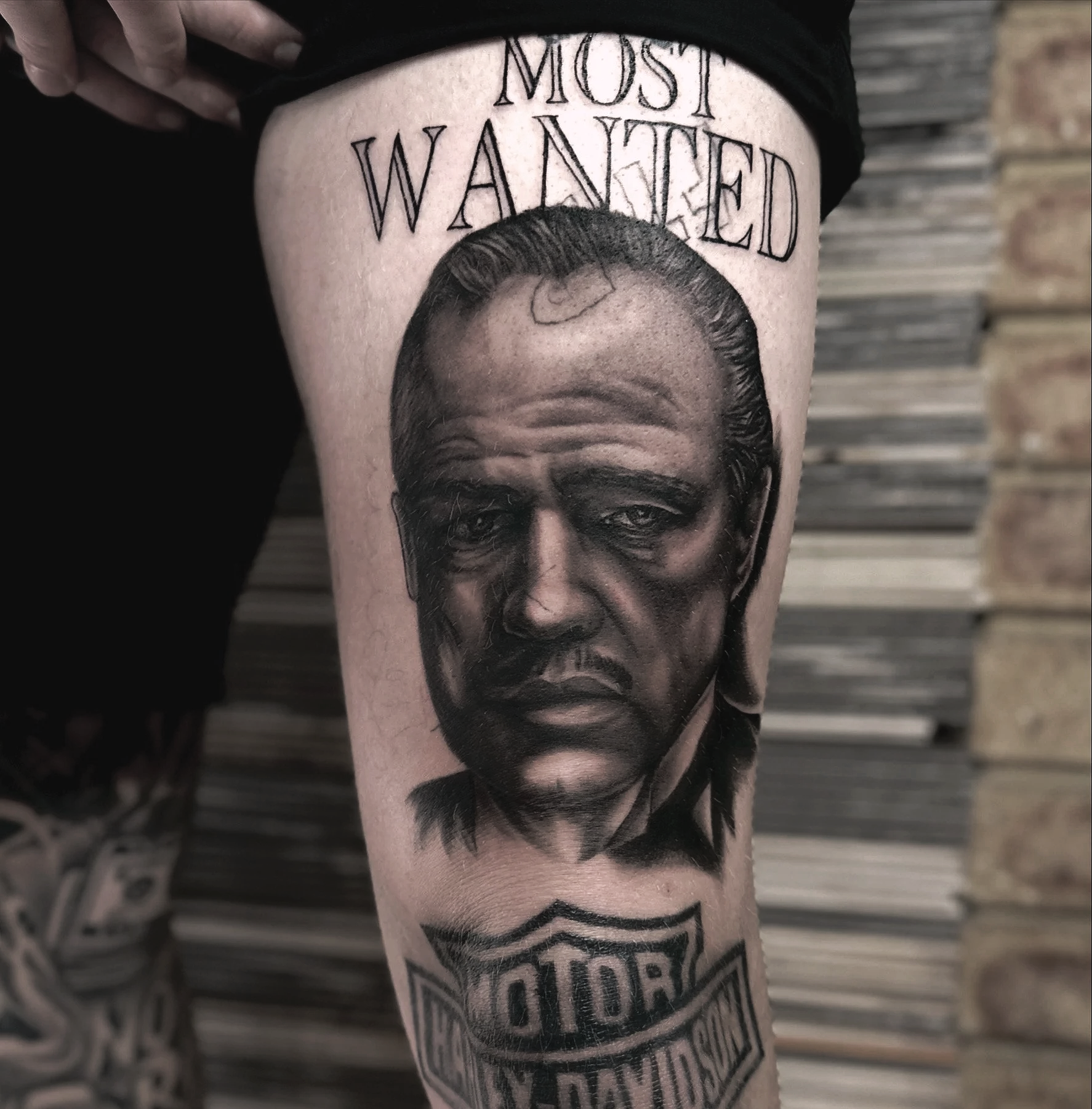 A tattoo on a person's leg depicting a serious-looking man with slicked-back hair, above the phrase 'Most Wanted', and below a Harley-Davidson logo with the words 'Harley-Davidson'.