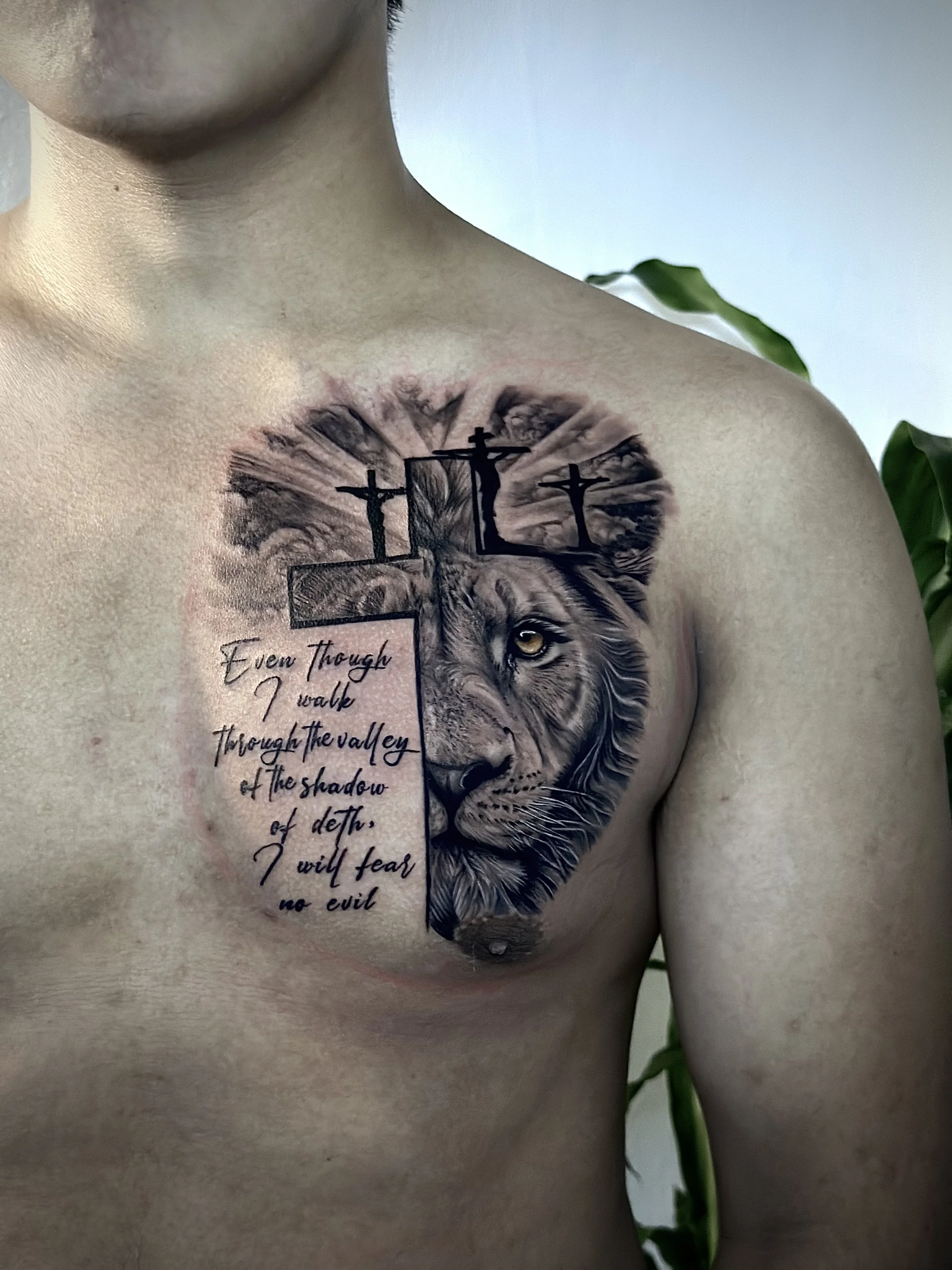 Tattoo of a lion's face with a quote on the chest, crosses on the shoulders, and a background of a cloudy sky.