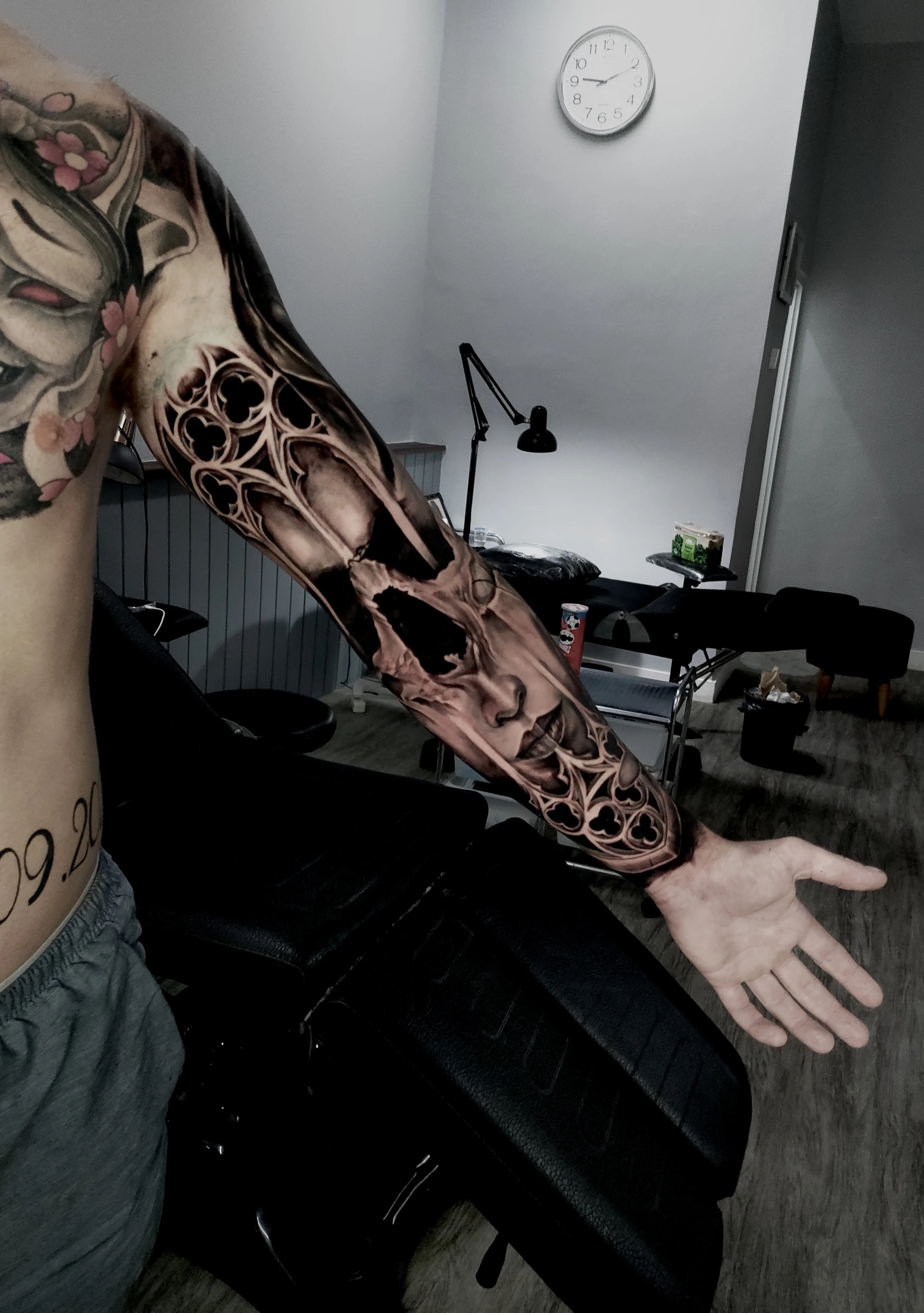 Tattooed arm extended in a room with a clock on the wall, a black desk lamp, and furniture.