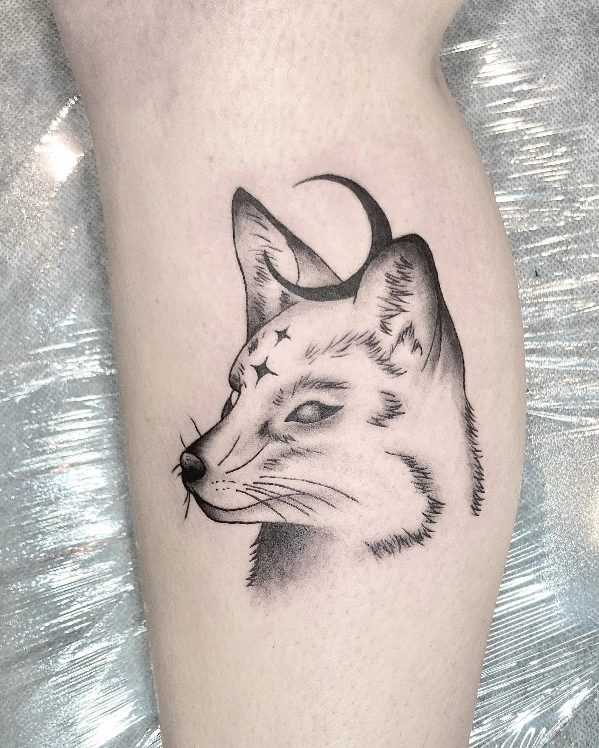 Black and gray tattoo of a fox's head with a crescent moon on its forehead, on person's skin.