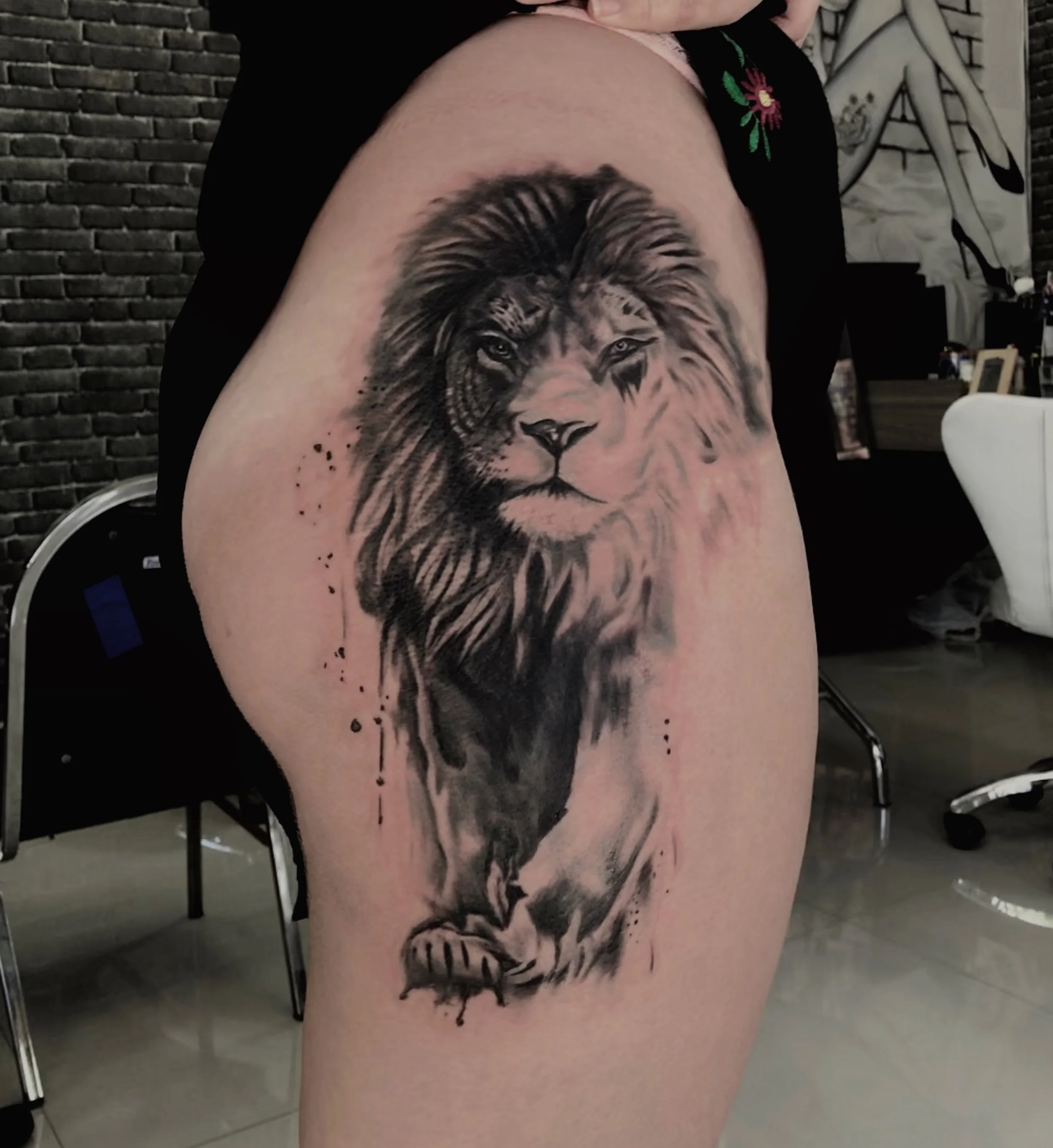 A black and gray tattoo of a lion's face and front paw on a person's upper thigh, with a watercolor and splatter style.