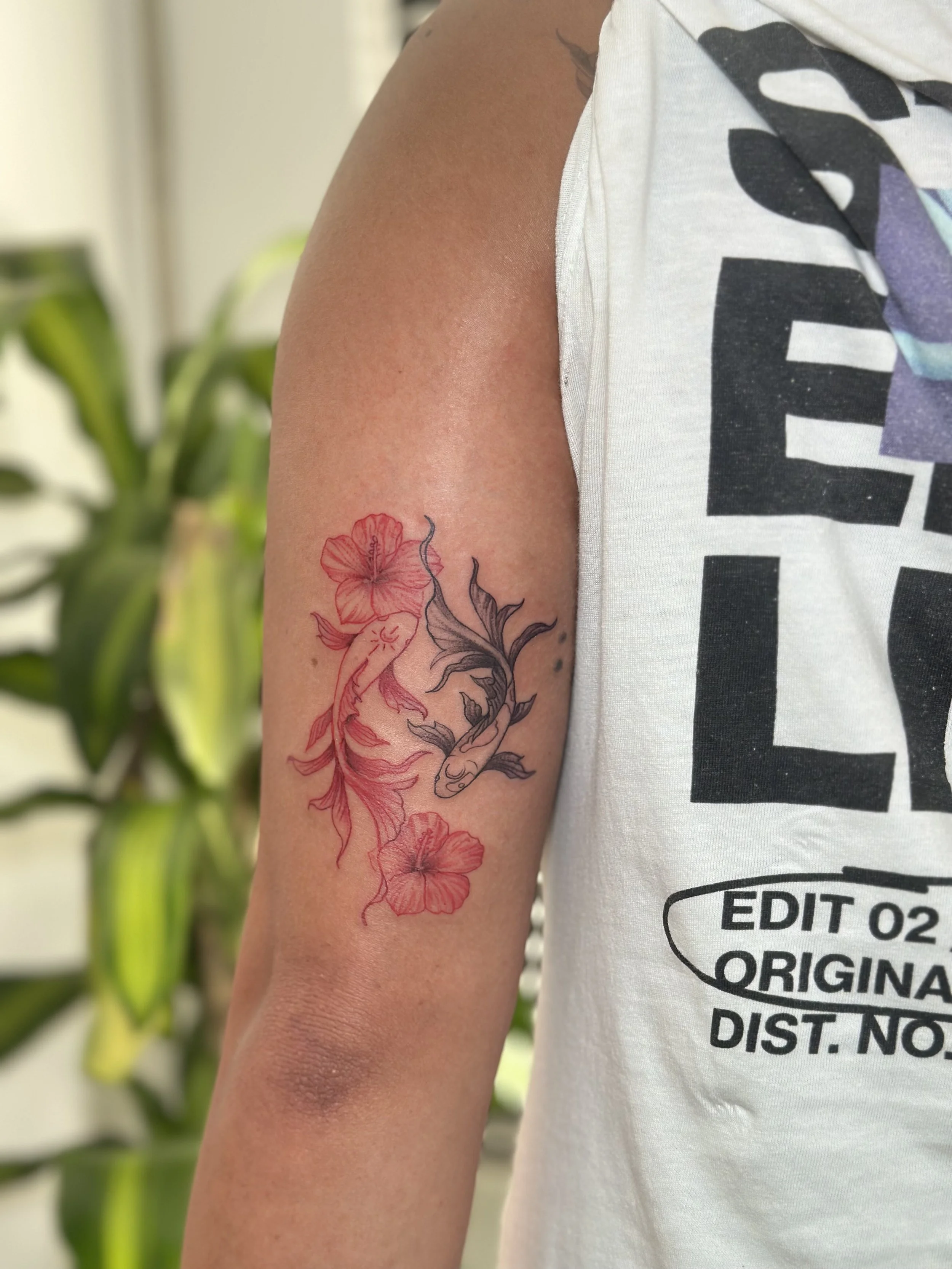 A person's arm with a colorful tattoo of a hibiscus flower and other floral elements, partially covered by a t-shirt with black text.