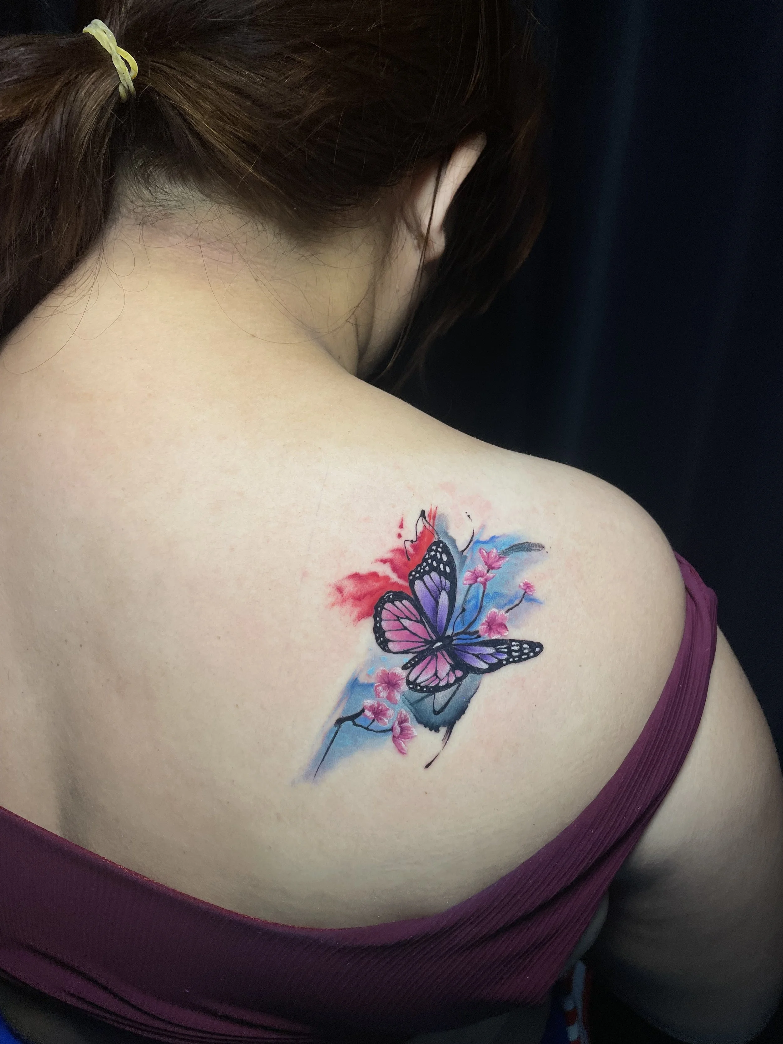 Tattoo of a purple and pink butterfly with pink flowers and blue background on a person's shoulder