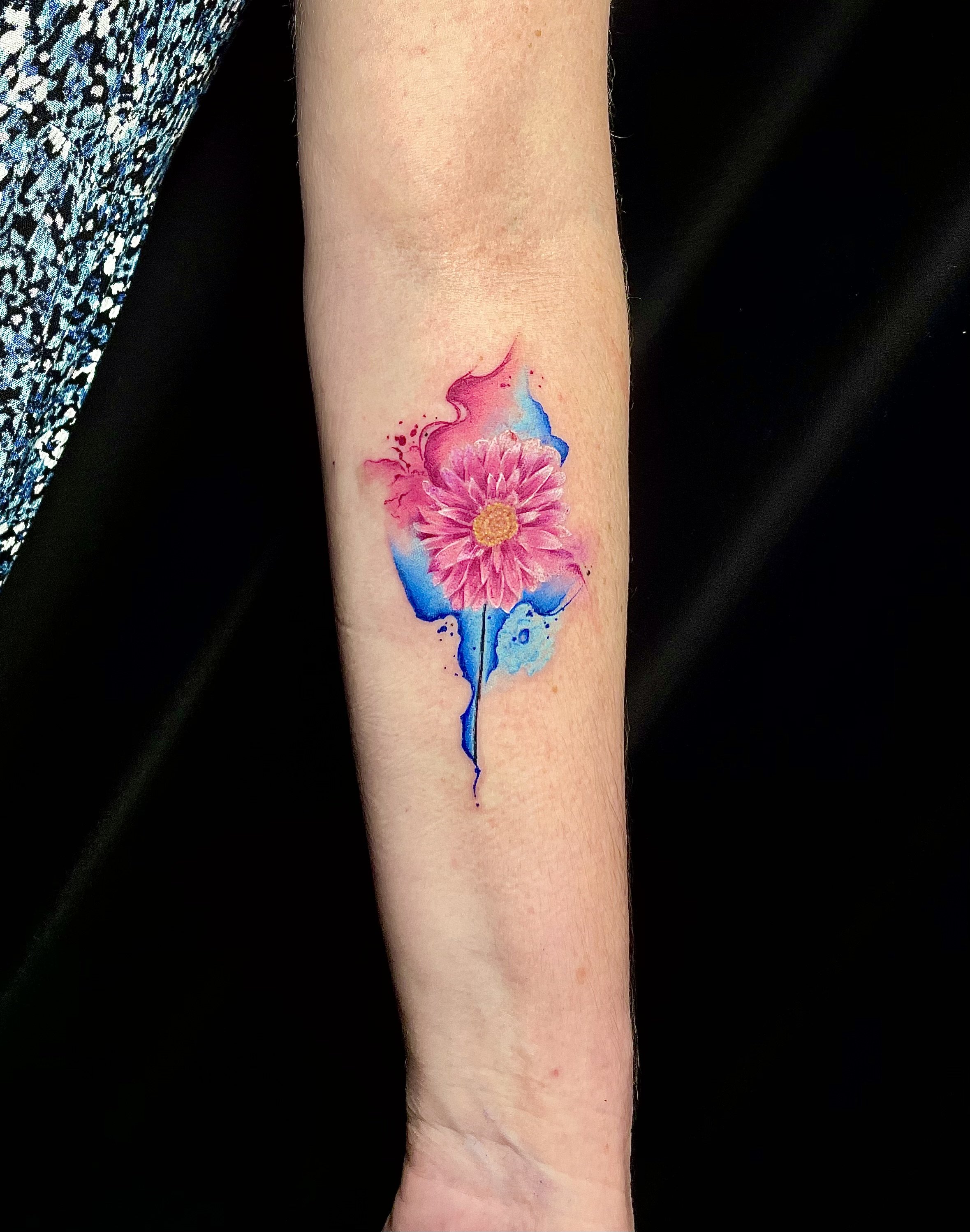 Colorful watercolor-style tattoo of a pink flower with a yellow center, with blue and pink splashes and drips around it on a person's forearm.