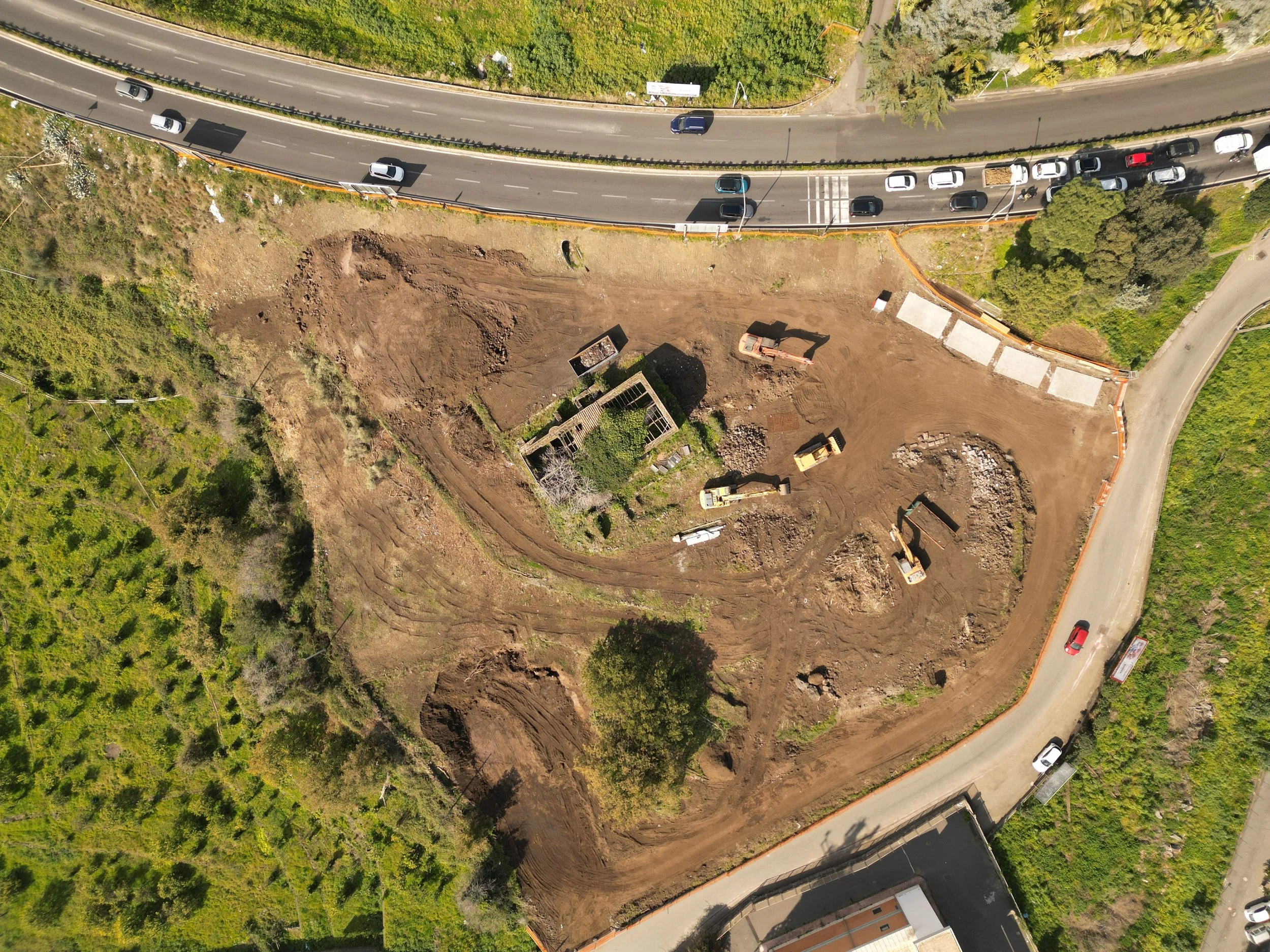 Area cantiere Spazio Umano Acireale Immagine aerea del cantiere Spazio Umano con escavatore e terra smossa, accanto a una strada trafficata con auto parcheggiate e in movimento, in un'area ricca di vegetazione e alberi.