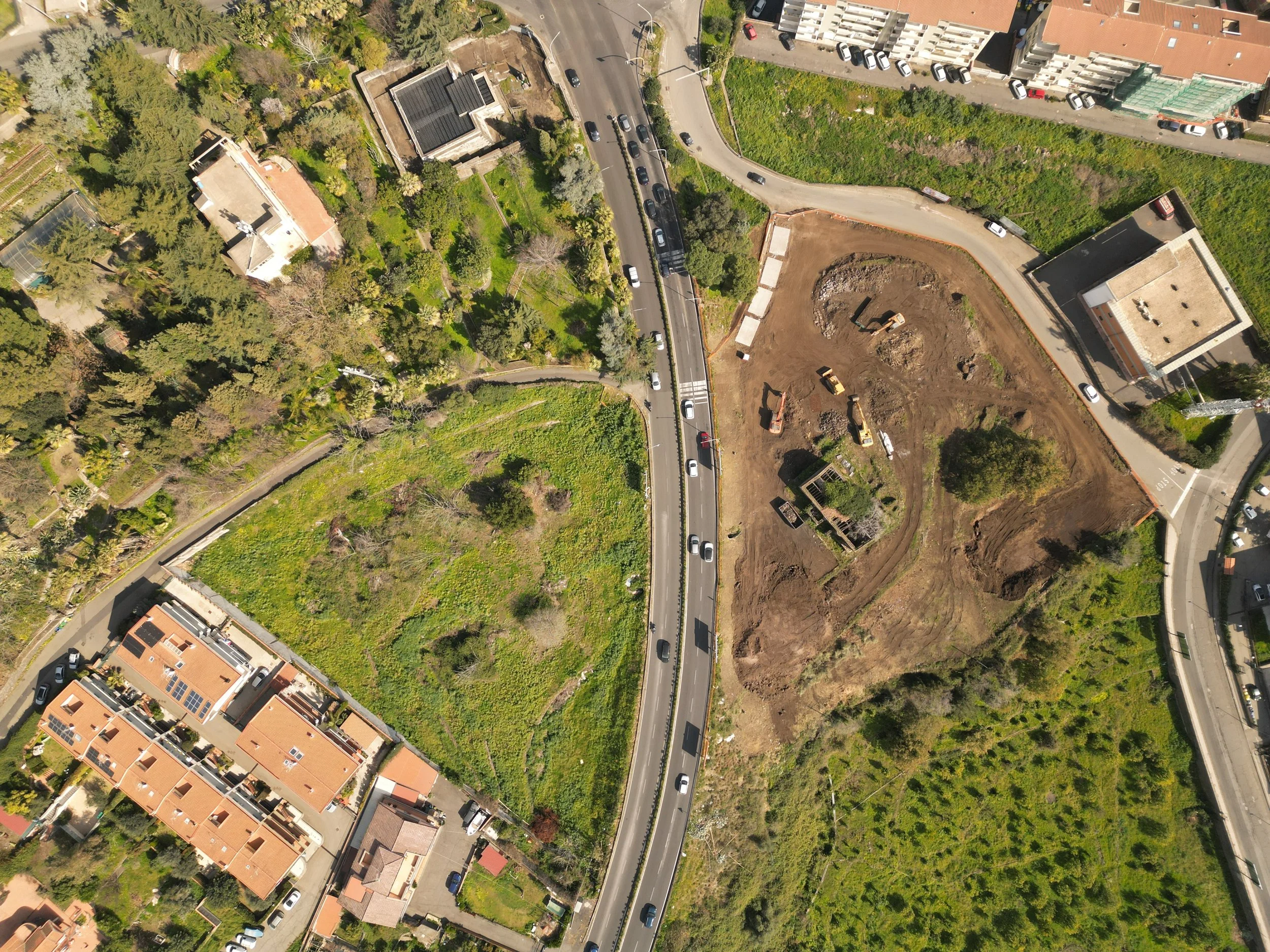 Area cantiere Spazio Umano Acireale  Veduta aerea di una strada in fase di costruzione circondata da aree verdi e case. Sono presenti veicoli parcheggiati e macchinari edili.