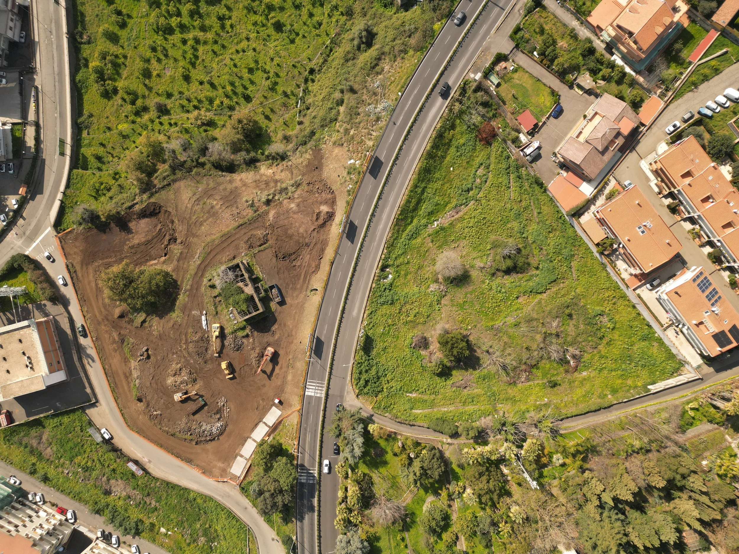 Acireale Veduta aerea di una strada che attraversa un'area urbana con edifici, parcheggi e spazi verdi, alcune parti sono in costruzione o in ristrutturazione.