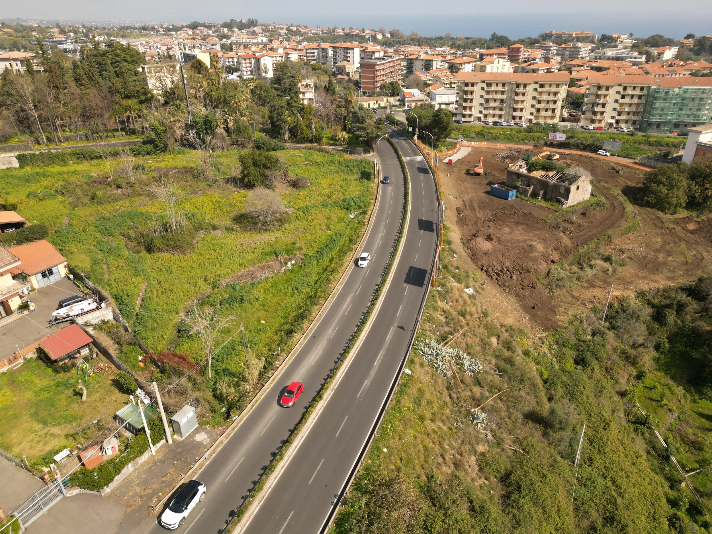 Area cantiere Spazio Umano Acireale  Vista aerea di una strada curva circondata da edifici e aree verdi, con un'area in costruzione e alcuni veicoli in movimento.