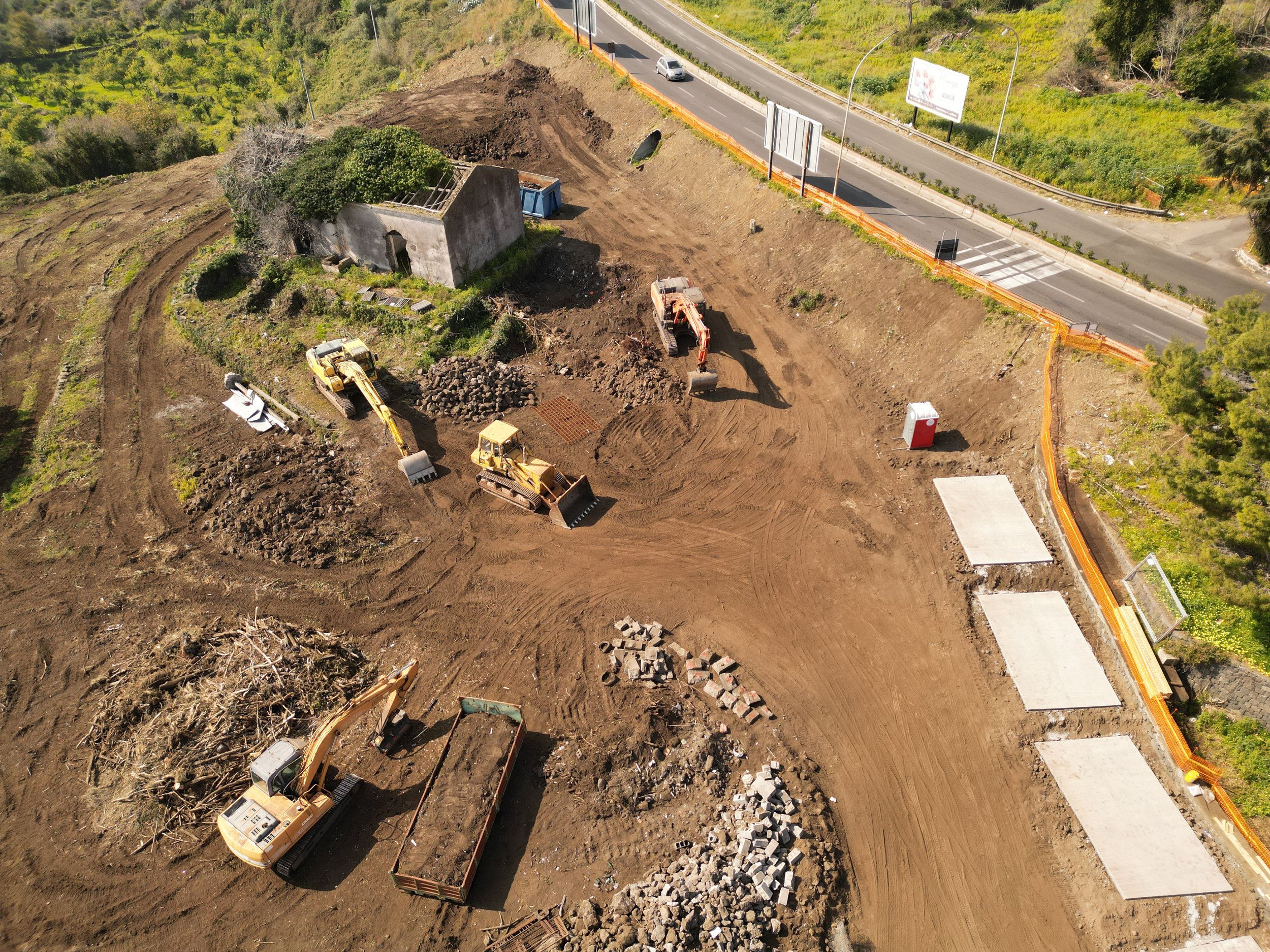 Area cantiere Spazio Umano Acireale  Cantiere di lavori stradali con escavatrice, mietitrebbia e altri macchinari, un edificio abbandonato e una strada sulla collina con cartelloni pubblicitari.