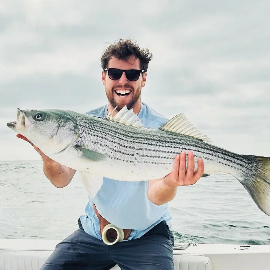 A man wearing sunglasses smiling and holding a large striped bass fish on a boat in the water.